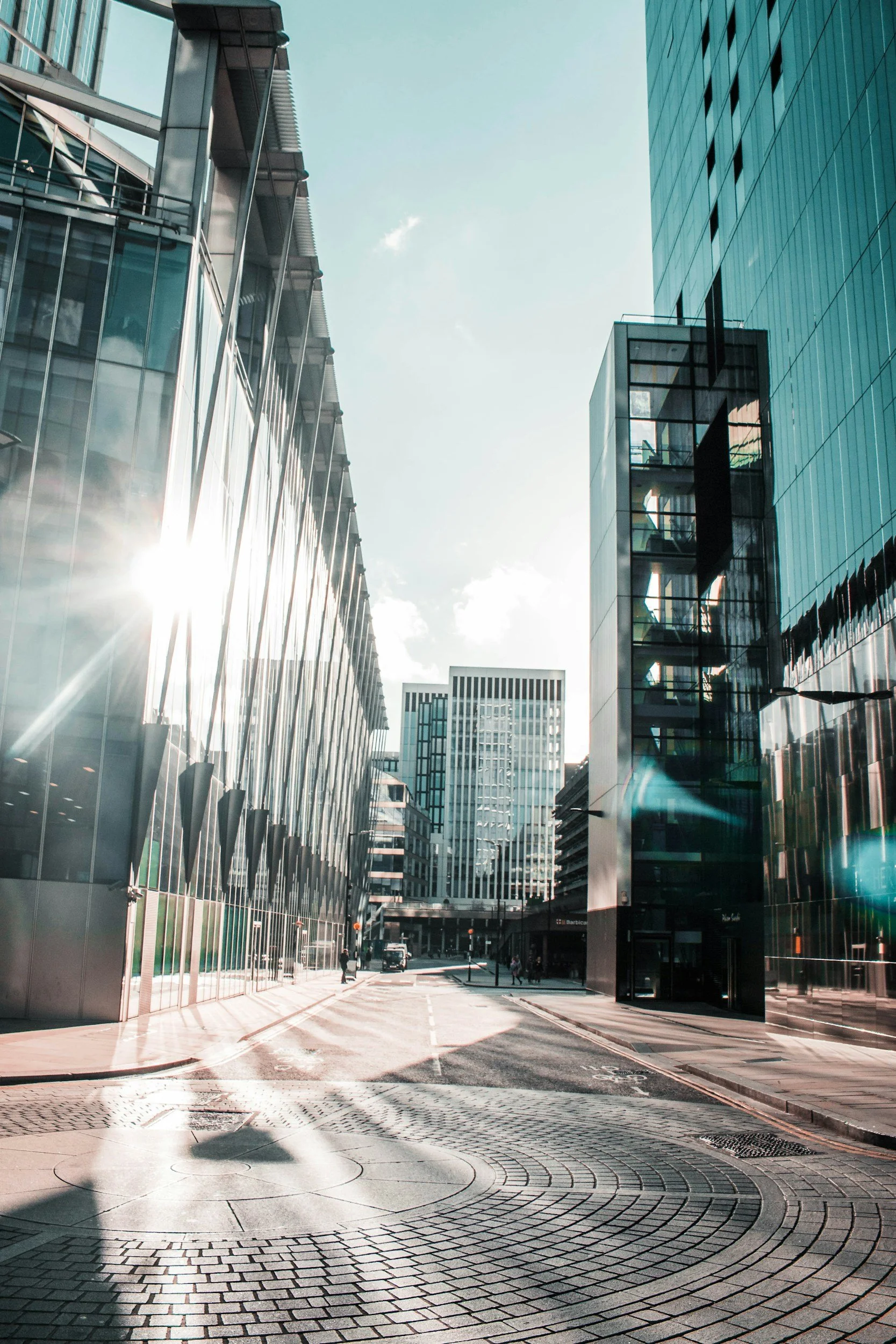 A city street surrounded by modern glass buildings with sunlight reflecting off the windows and a few pedestrians walking.