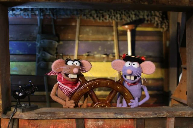 Two puppet mice dressed as sailors at a ship's helm in a wooden cabin.