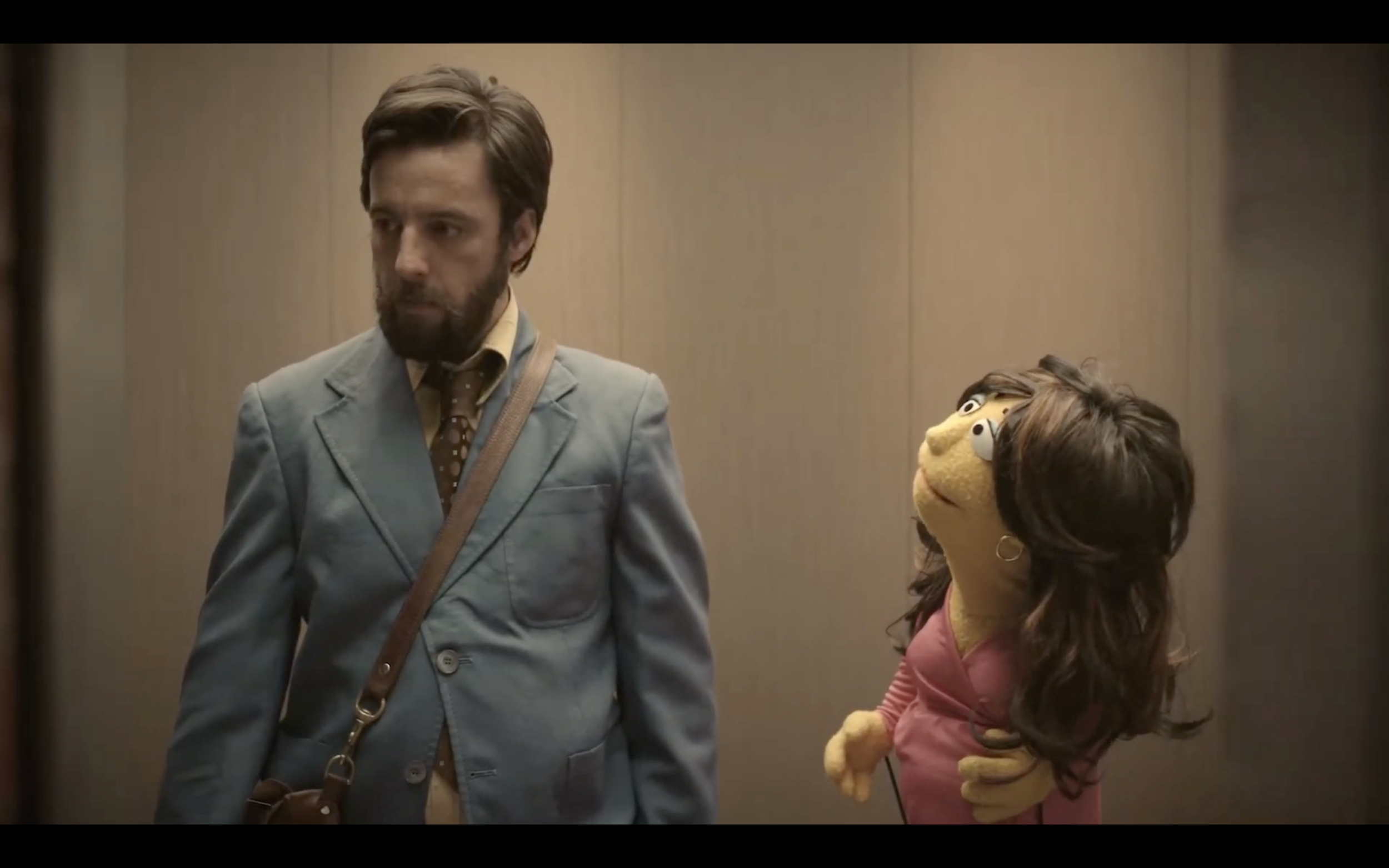 A man in a blue suit with a brown bag looks downward with a serious expression while a puppet with long brown hair, a pink dress, and large googly eyes looks up at him, standing in an elevator with wood-paneled walls.