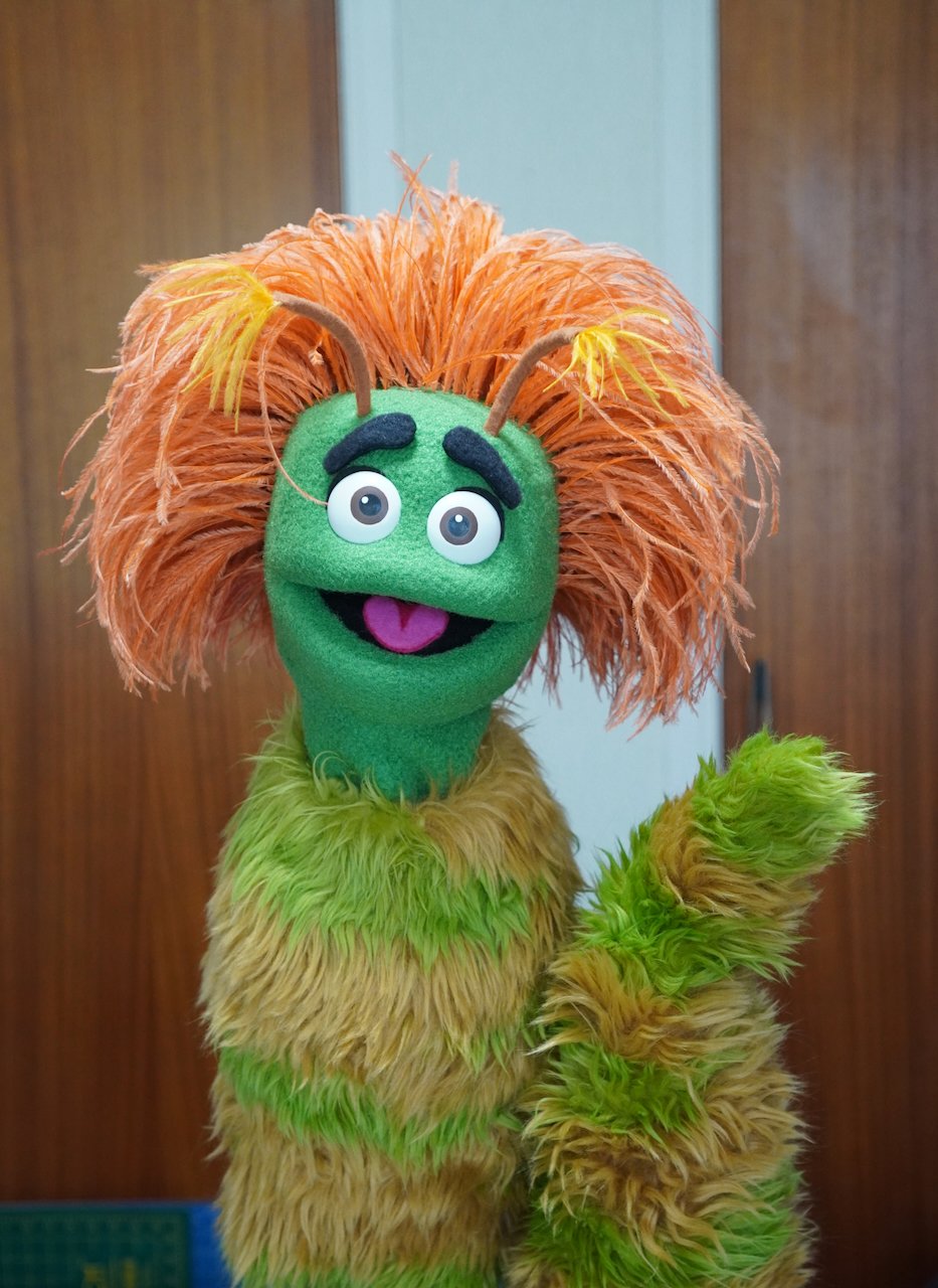 A colorful puppet character with orange, green, and brown furry features, large expressive eyes, thick black eyebrows, and antennae on its head, standing against a wooden background.