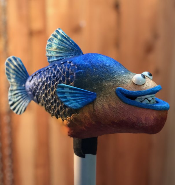A fish puppet with a wide toothy smile, mounted on a stick, against a wooden fence background.
