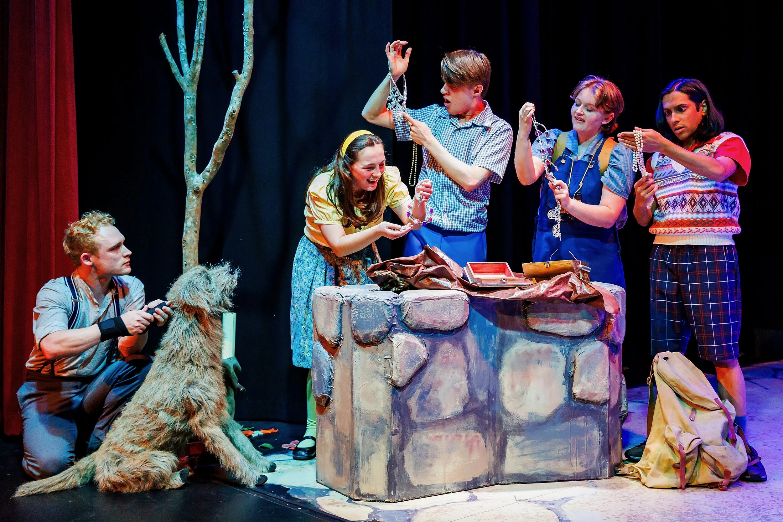 A group of young actors performing on stage, with some holding jewelry and a person with a dog puppet, set against a dark background with a painted stone-looking surface and a tree prop.