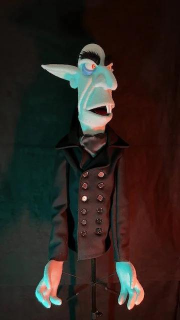 A puppet with exaggerated facial features and light blue skin, dressed in a black coat, standing against a dark background with colorful lighting.