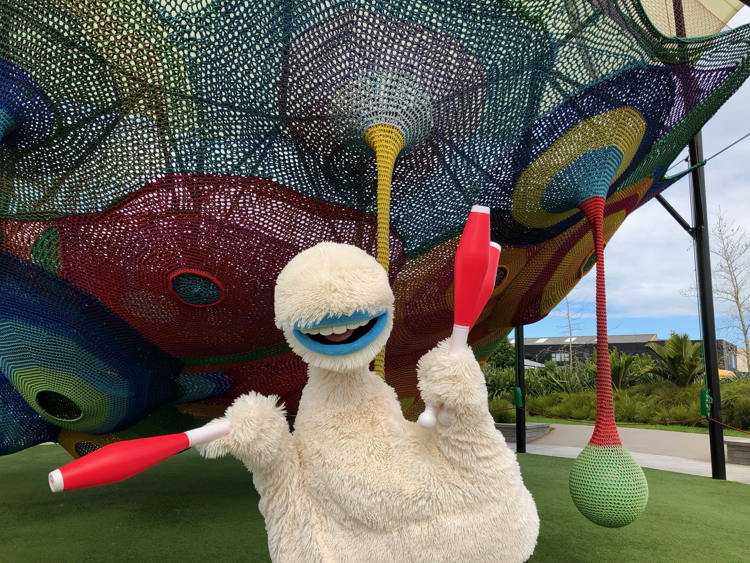 A giant furry puppet with a big smile and blue lips, holding two red and white juggling pins, standing in front of a colorful, woven outdoor art installation with various circular and abstract patterns.