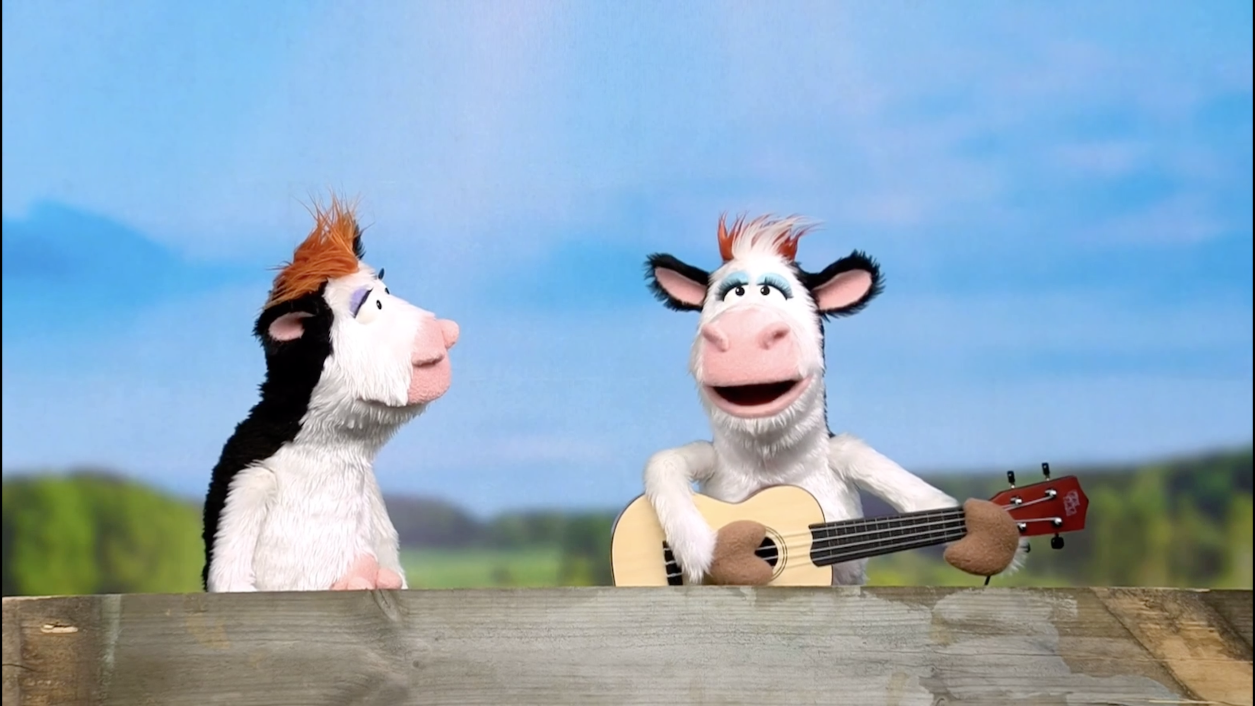 Two puppet animals with colorful fur, one holding a guitar, performing outdoors with a blue sky and green landscape in the background.