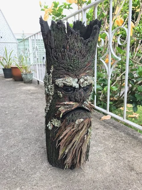 A tree trunk carved and painted to resemble a fantasy character with a stern face, eyebrows, eyes, and a large beard, placed on a rooftop terrace with potted plants and a decorative white metal railing.