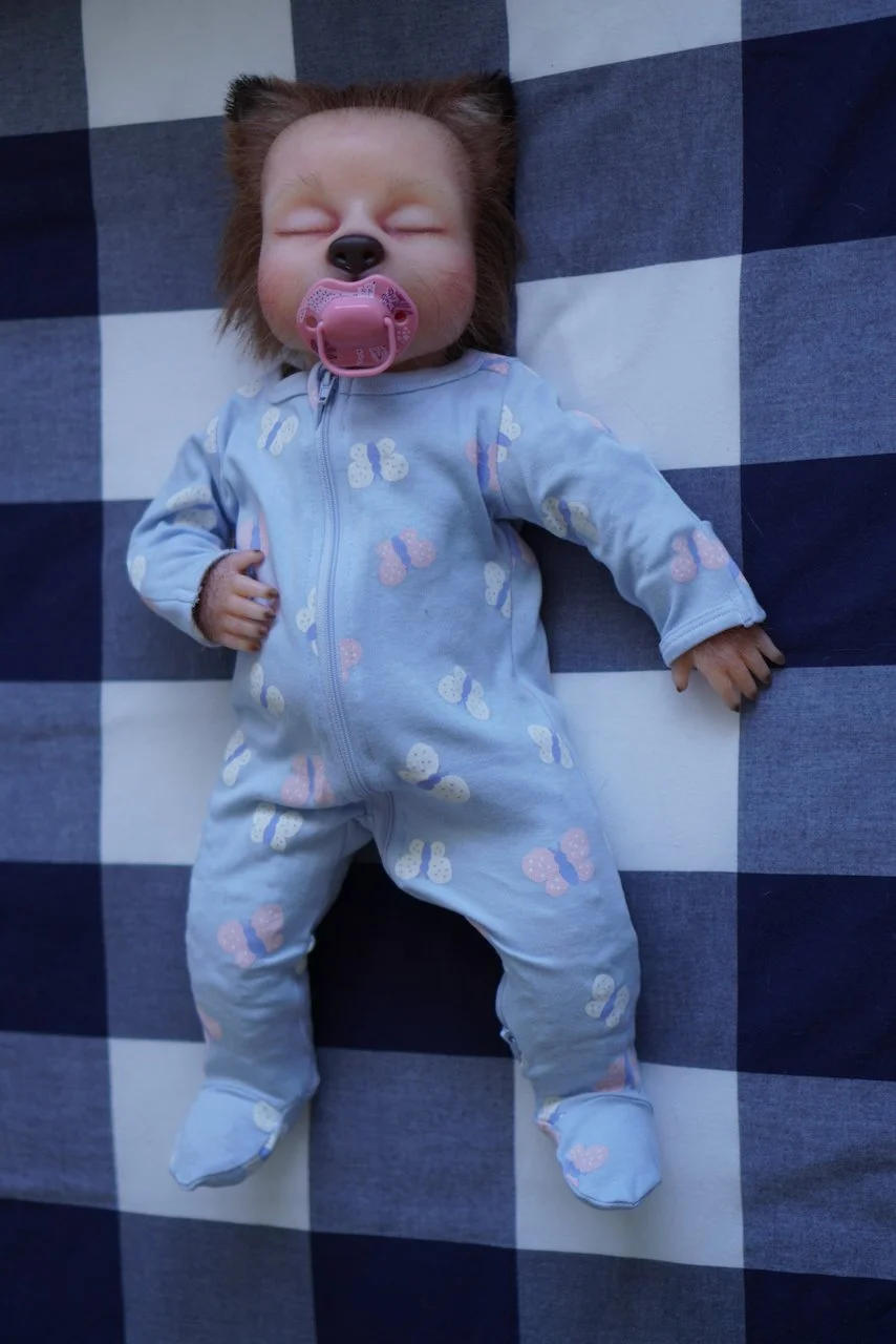 Baby fox child puppet with a pacifier, wearing a light blue onesie with butterfly prints, lying on a black and white checkered surface.