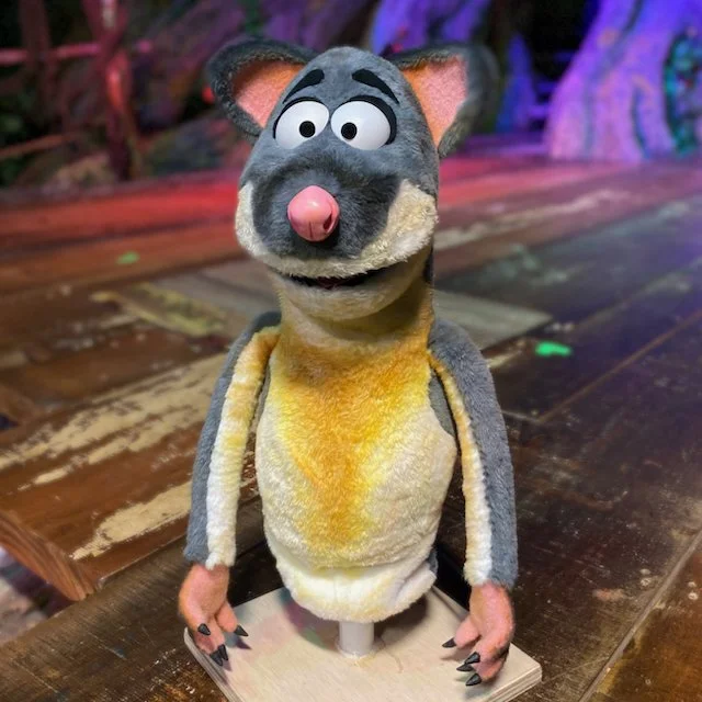A puppet of a possum with a gray face, big white eyes, a pink nose, and a wide mouth, sitting on a wooden table overall with a colorful background.