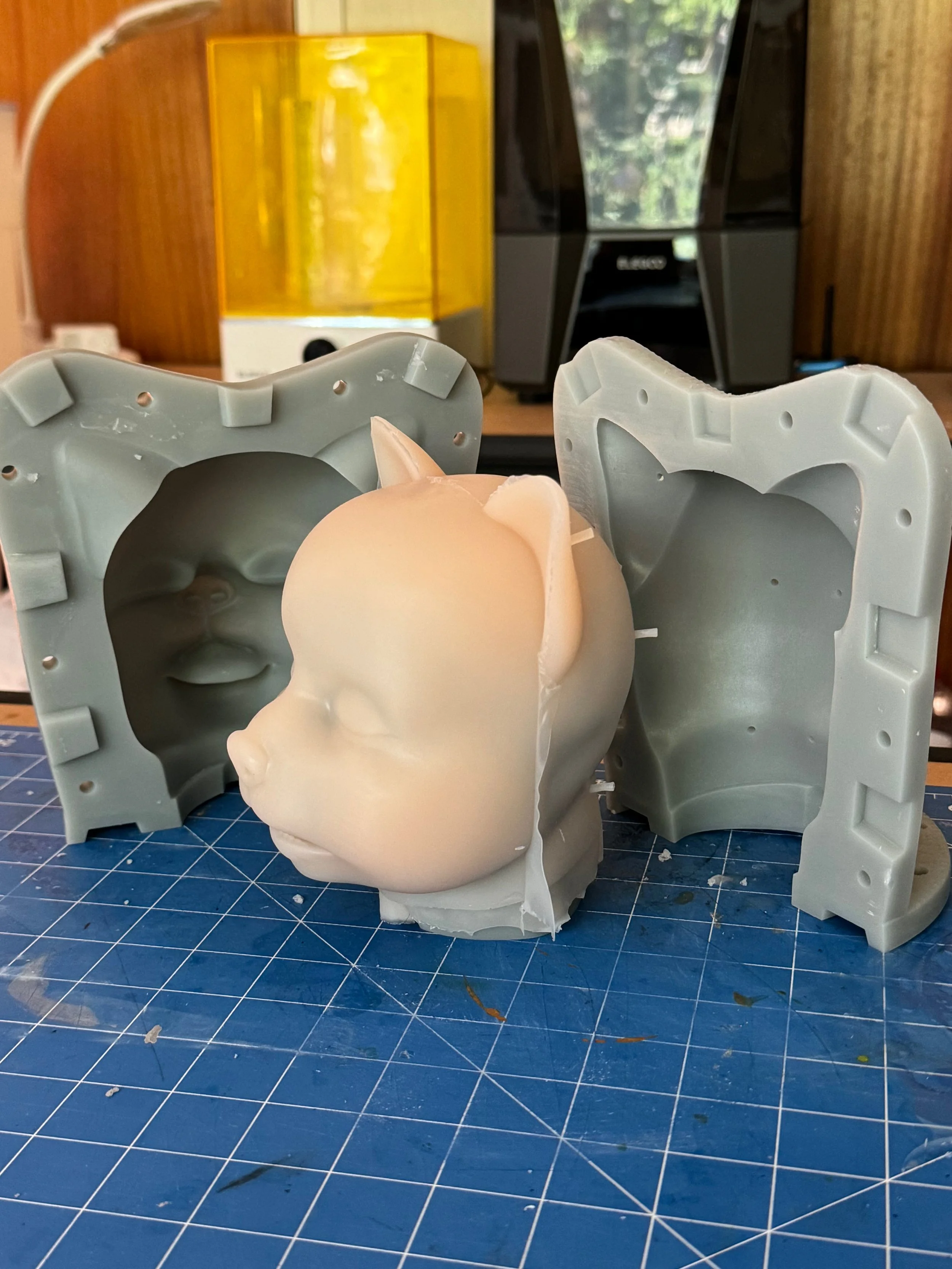 3D-printed mold and a silicone prototype of a girl's face with a cat ear, placed on a blue cutting mat inside a workspace.