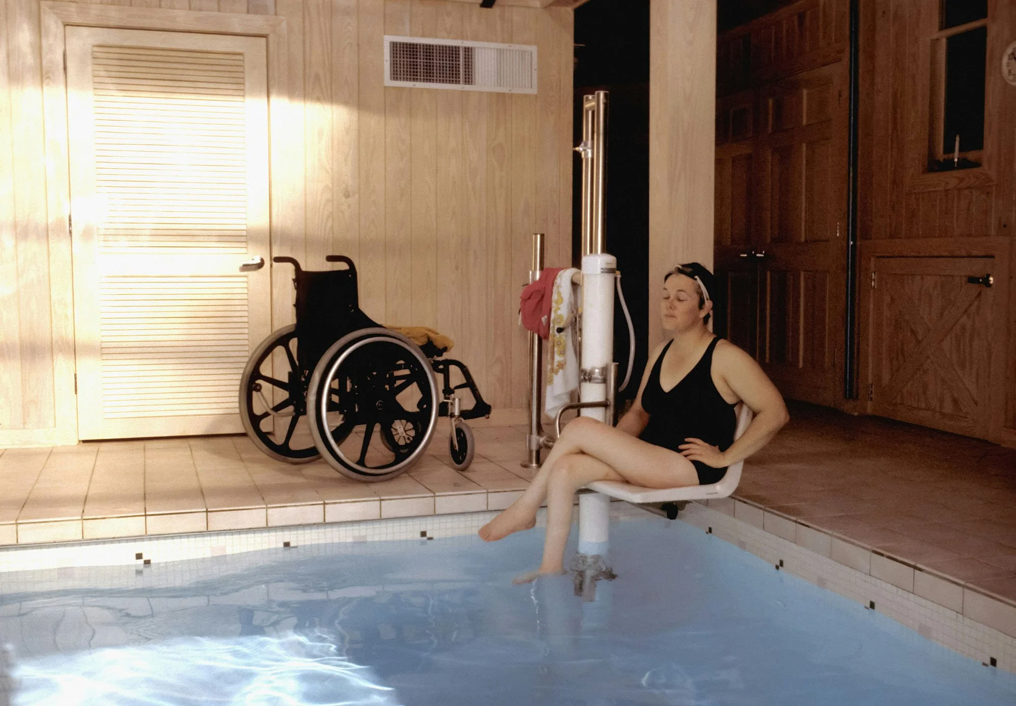 Woman sits on a hydro chair lift above water next to wheelchair for hydrotherapy session
