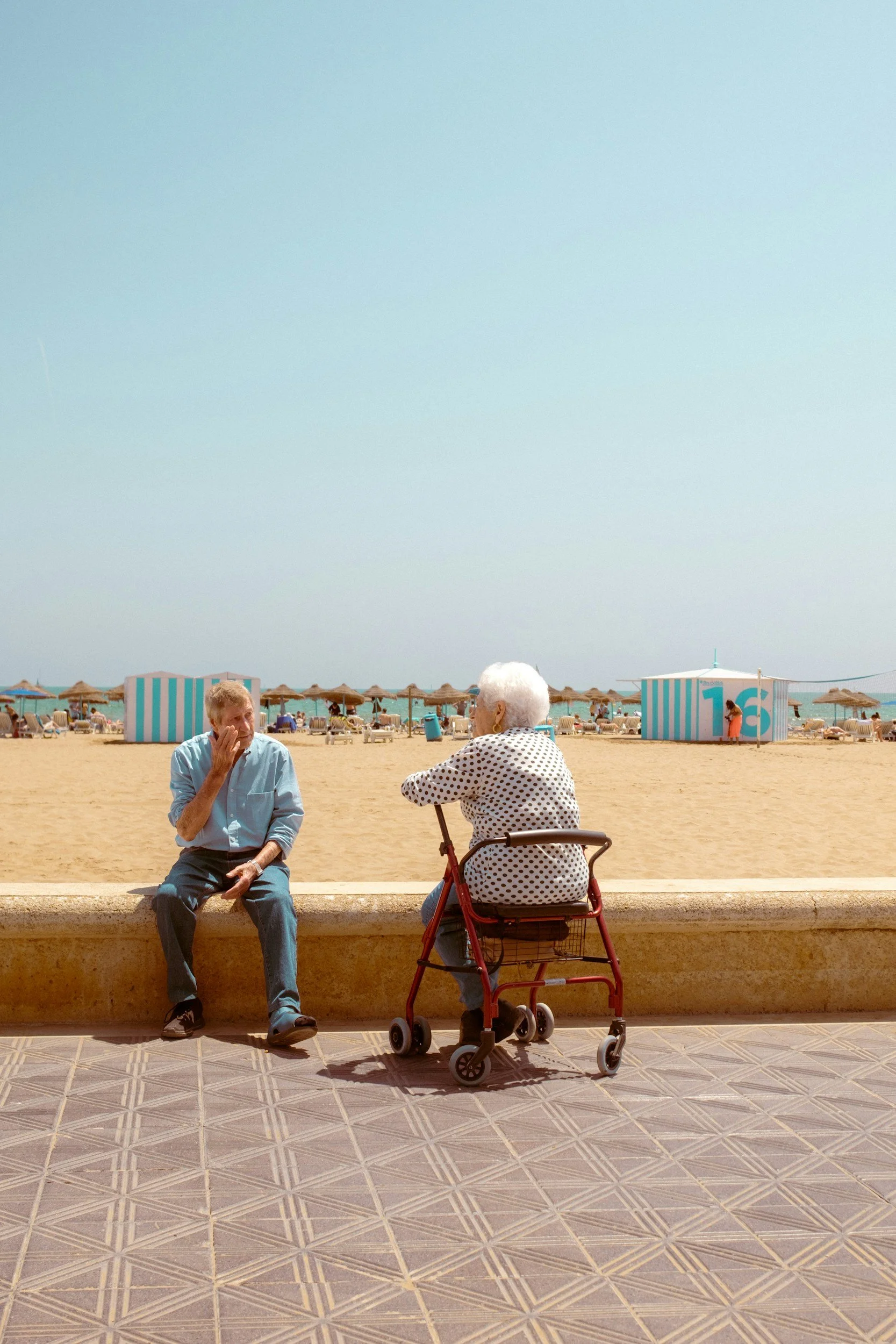 An elderly woman with white hair sitting on a walker talking to a man on a bench at the beach, with beach umbrellas and two large striped beach huts in the background.