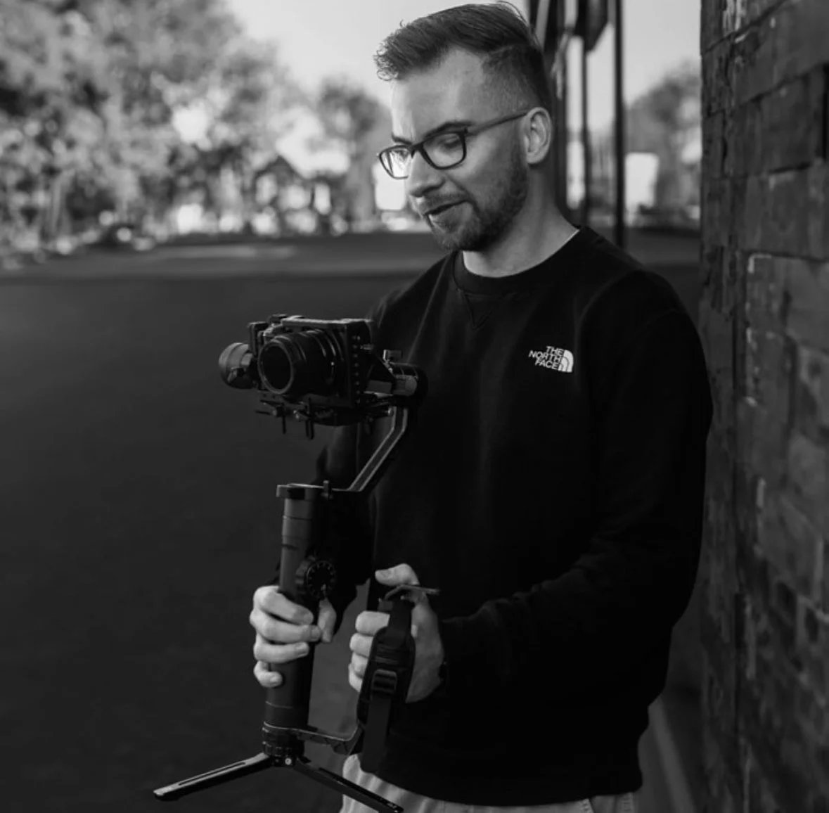 A man in glasses and a black North Face sweatshirt holding a camera mounted on a stabilizer against a brick wall, outdoors.