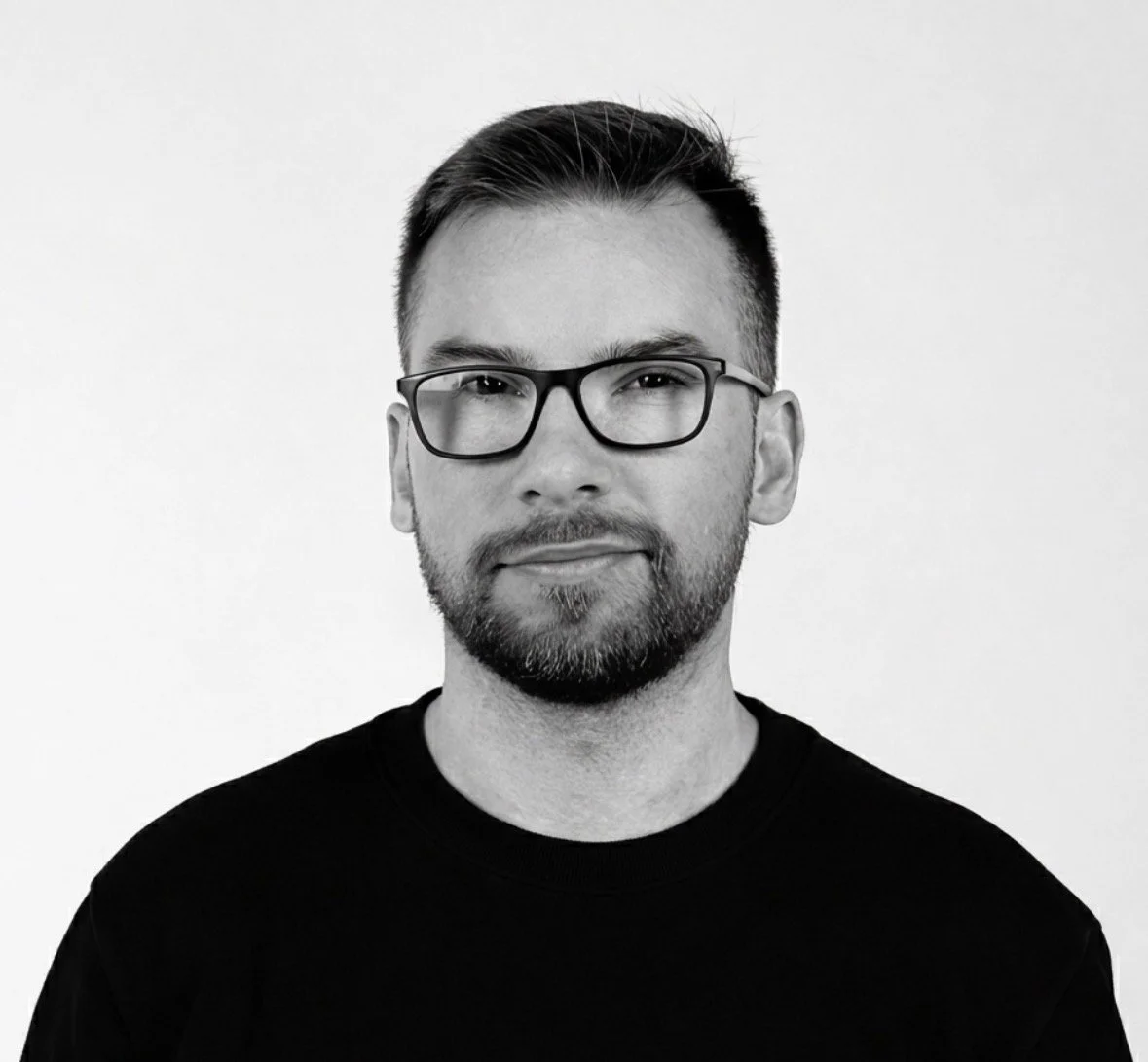 Black and white portrait of a man with glasses, short hair, and a beard, wearing a black shirt, looking directly at the camera.