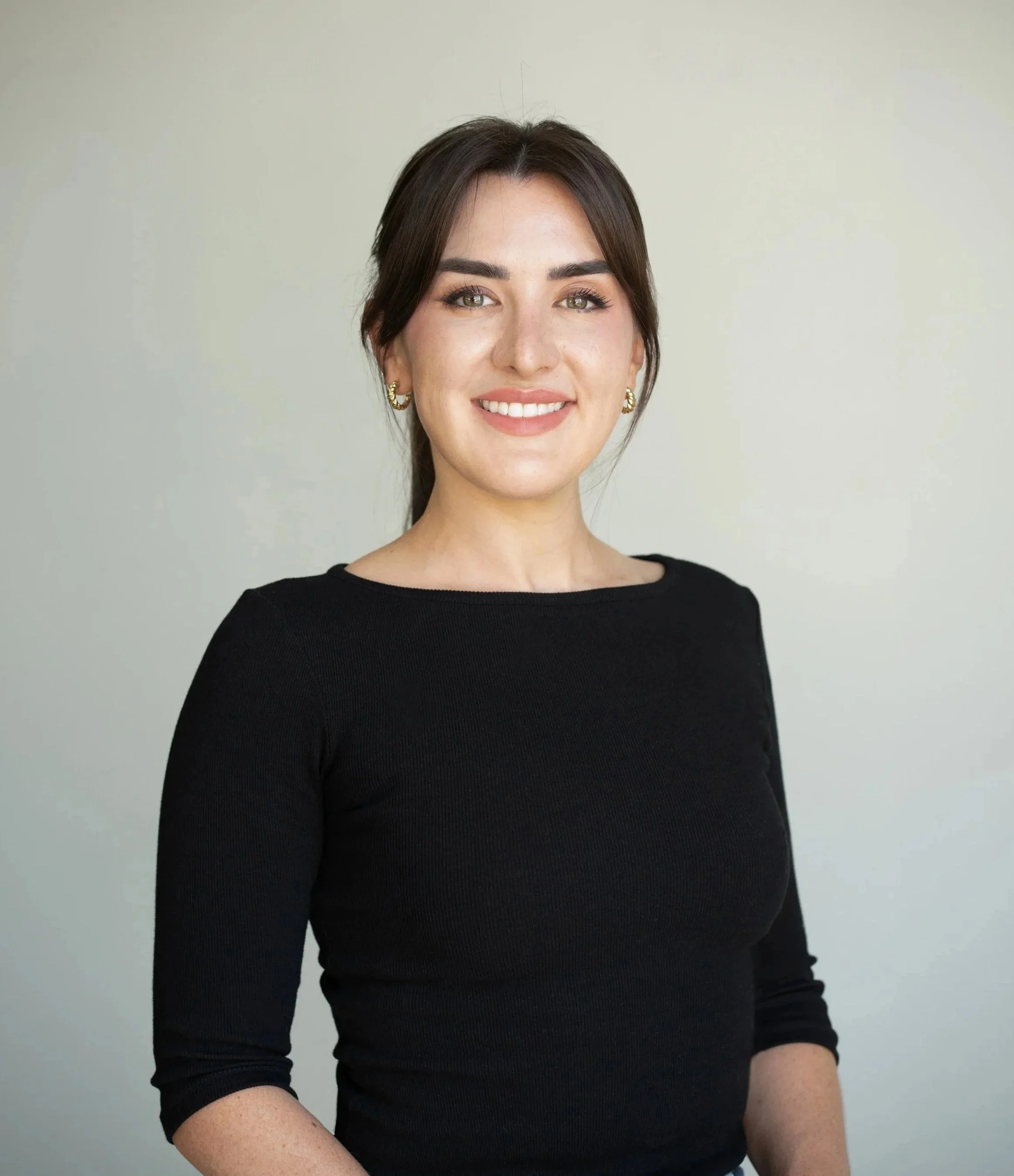 A woman with dark brown hair, light skin with freckles, green eyes, and wearing gold earrings and a black long-sleeve top, smiling at the camera against a plain light background.