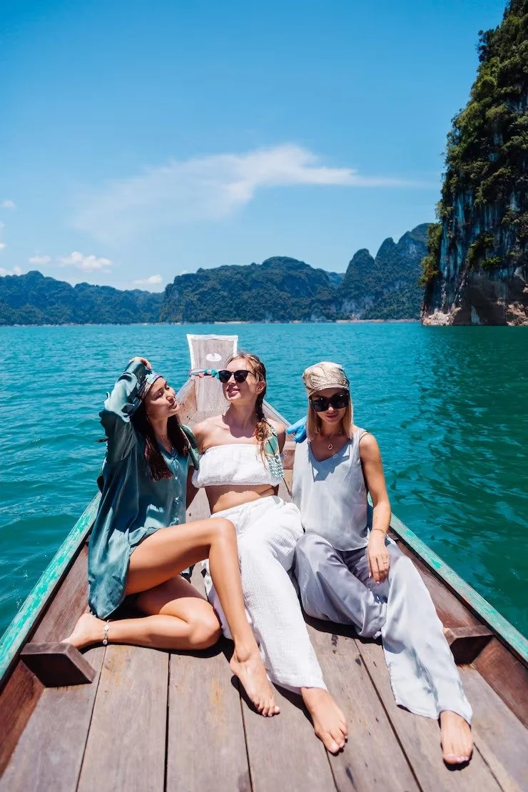 Three women sit on a wooden boat floating on turquoise water, with lush green cliffs and blue sky in the background.