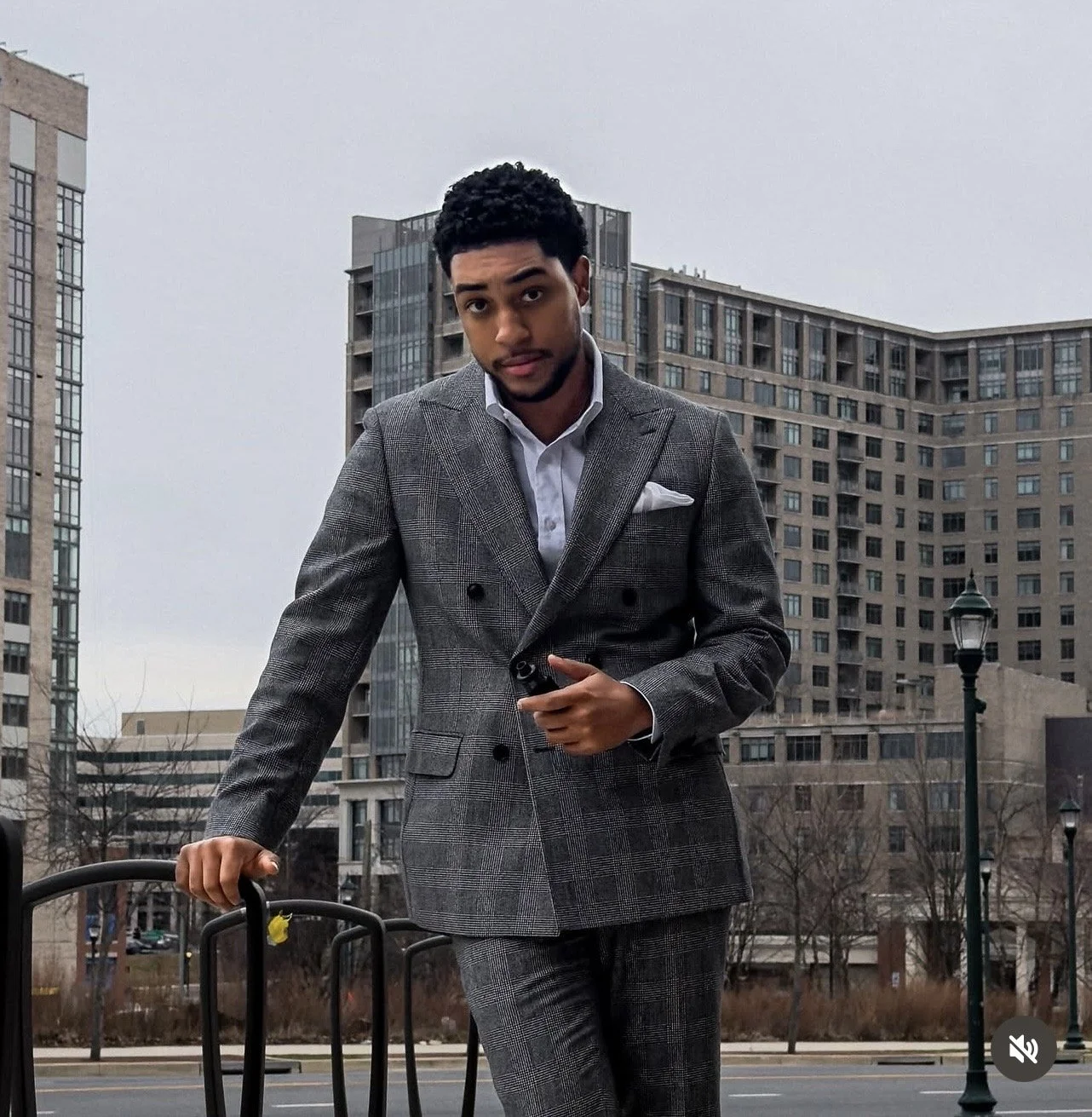 Young man in a gray plaid suit and white shirt standing outdoors in an urban area with tall office buildings and leafless trees in the background.