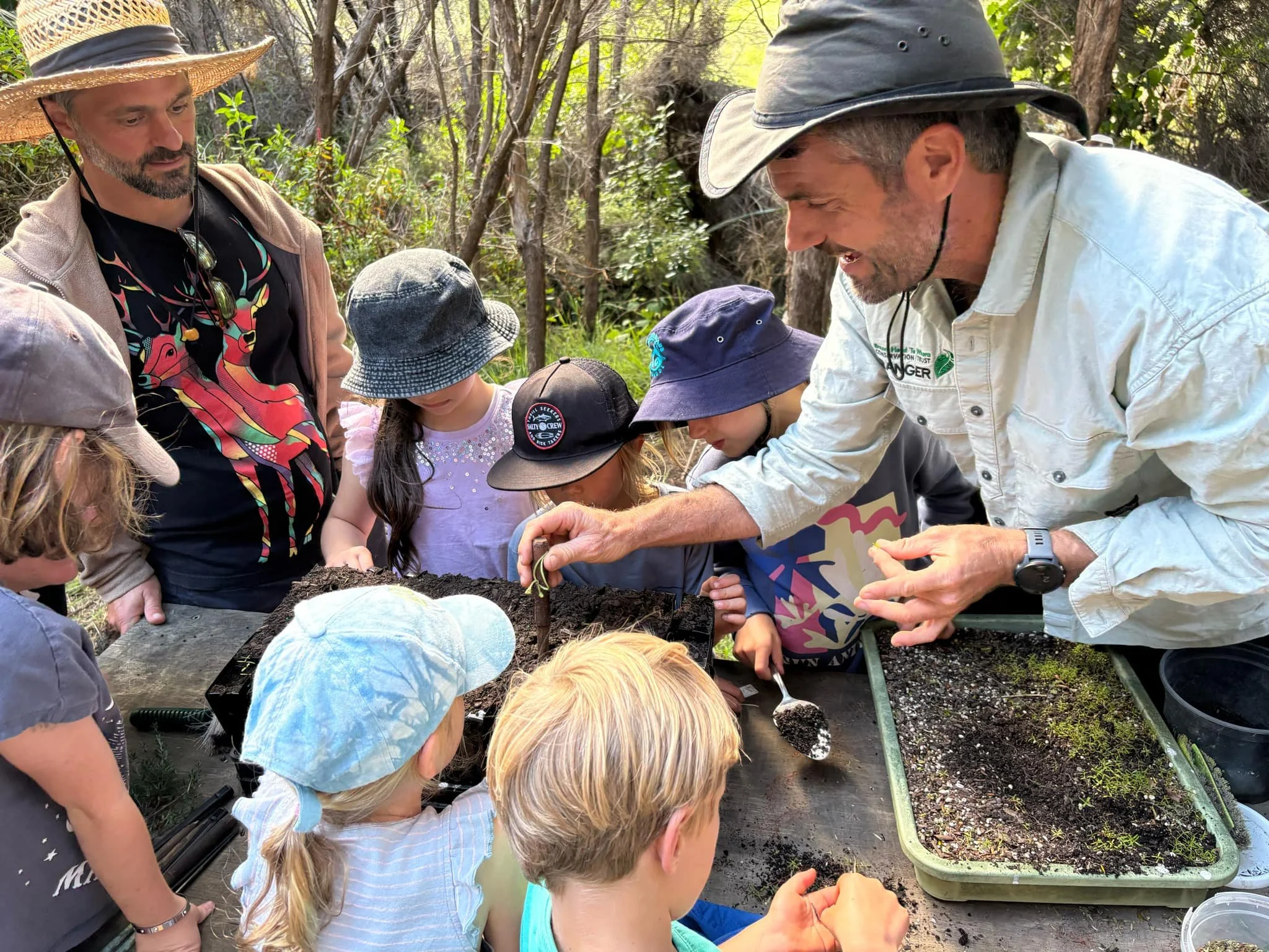 Group of children and two adults gathered around a table outdoors, examining soil or small plants, engaging in a gardening or educational activity in a wooded area.