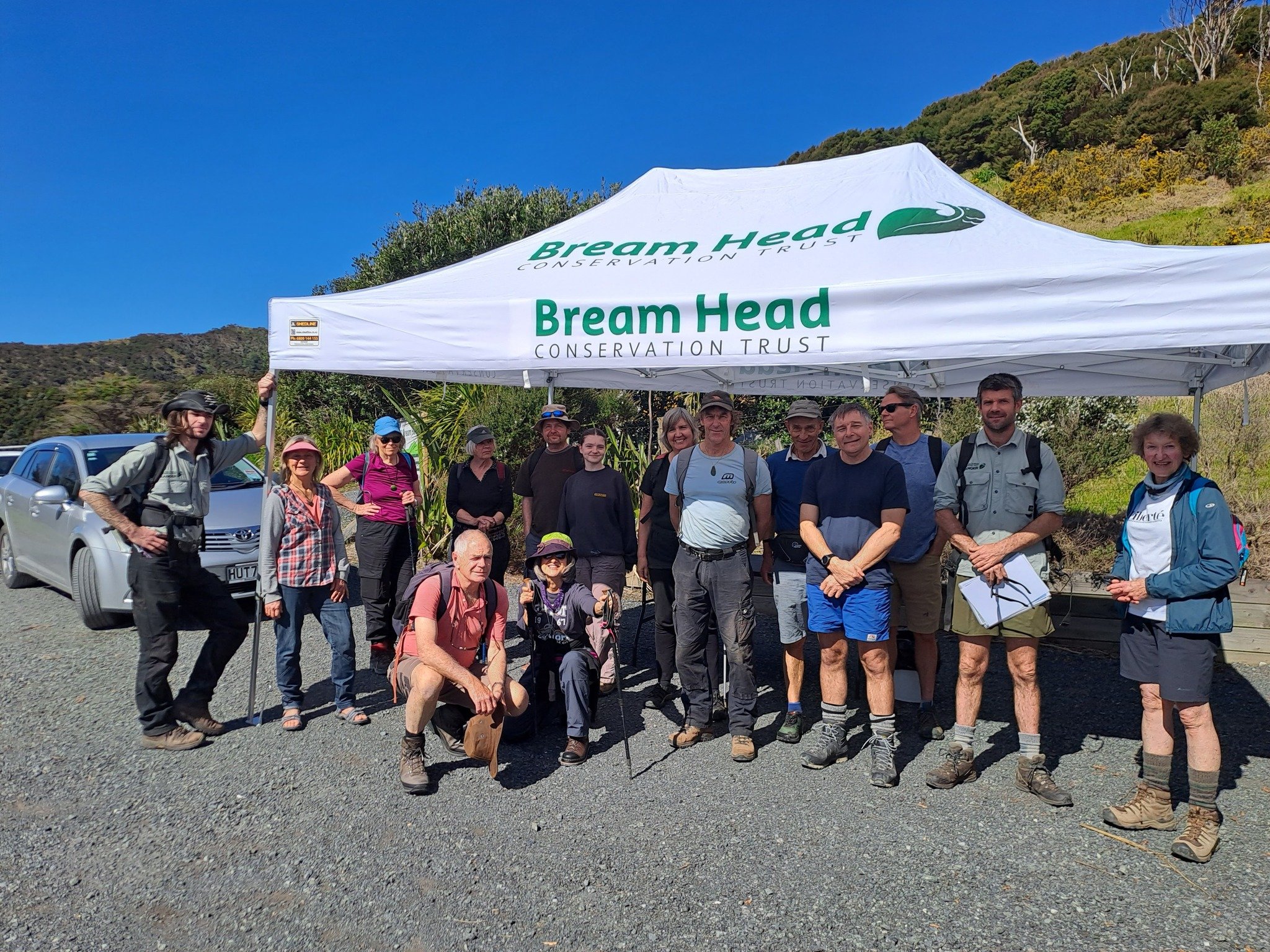 Group of people standing and kneeling outside in front of a white canopy tent with Bream Head Conservation Trust logo, in a natural setting with mountains and trees in the background, under a clear blue sky.