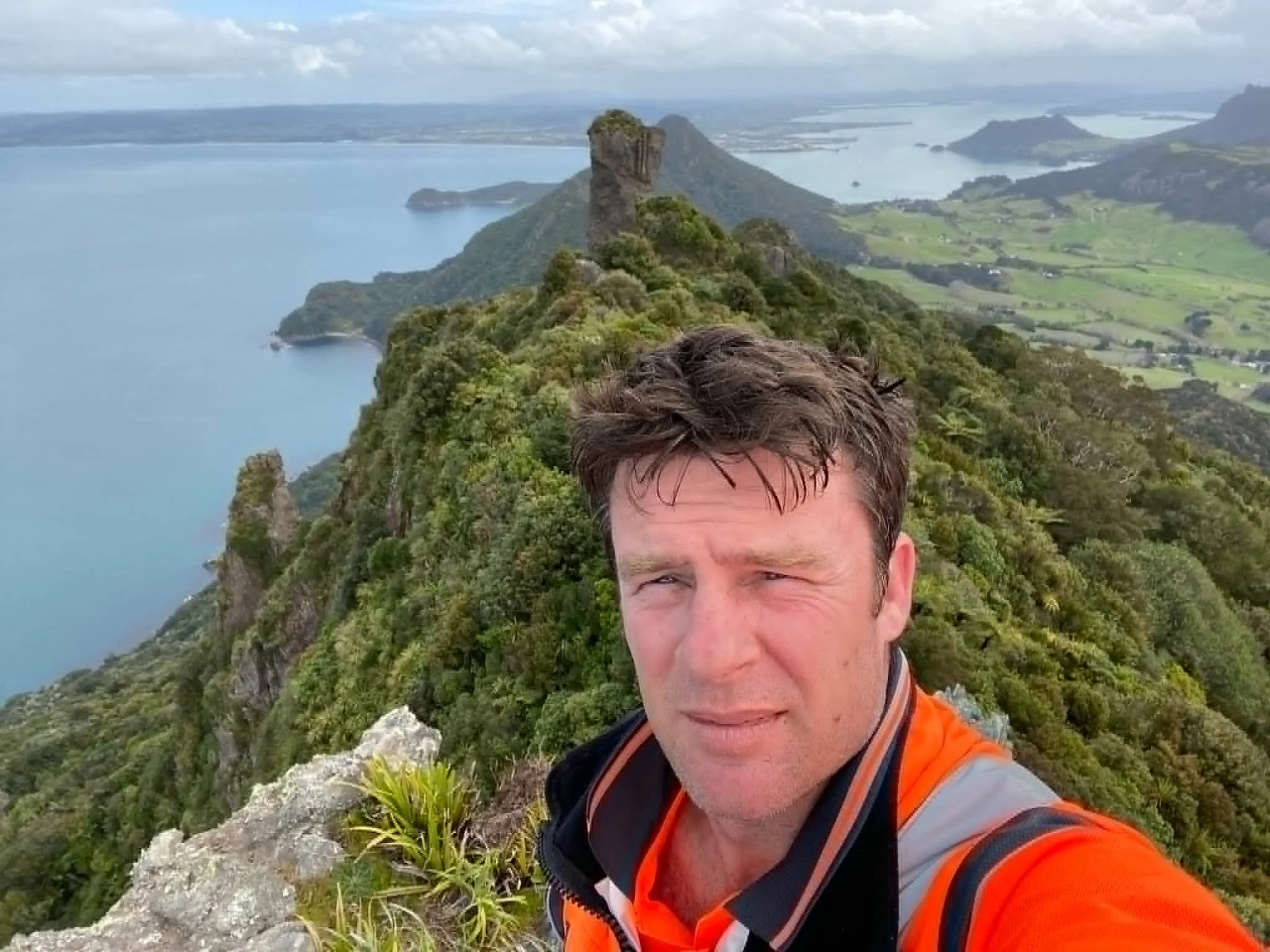 A man taking a selfie at the top of a mountain with a dramatic rocky peak and lush green landscape below, overlooking a body of water with islands and distant land.
