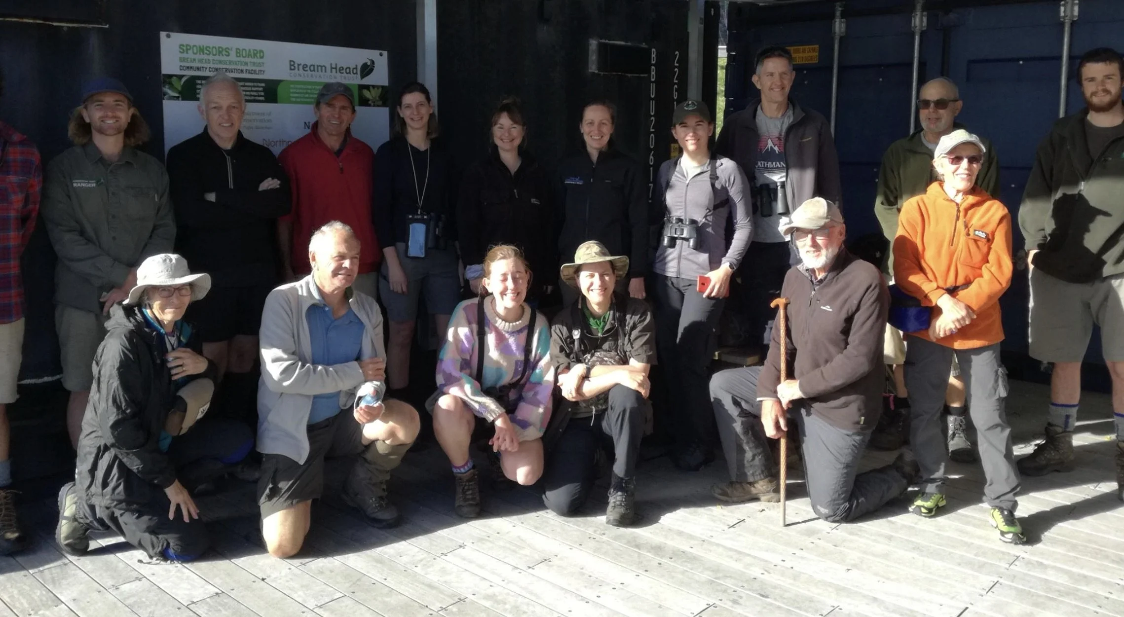 Group of people posed together outdoors, some holding hiking gear, under a sign for the Bream Head Conservation Trust.