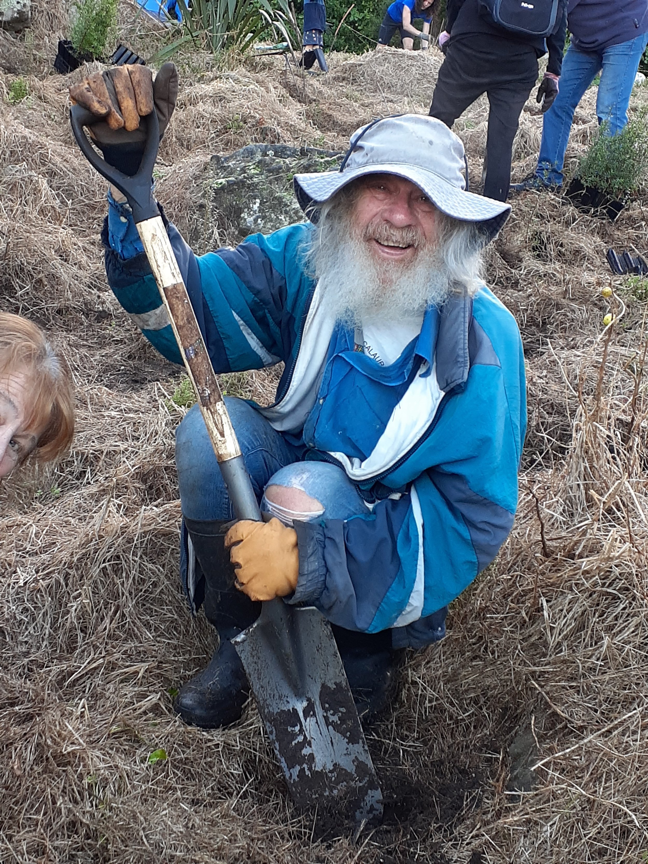 A man with a beard and long hair wearing a wide-brimmed hat, blue jacket, and yellow gloves kneels on the ground holding a shovel, smiling at the camera. He is in a dry, grassy outdoor area with other people working in the background, some with glove