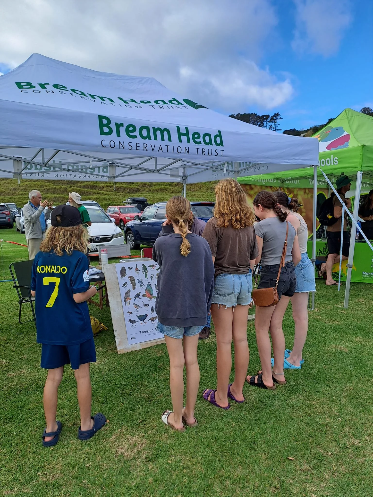 People gathered around an information booth at an outdoor event, with tents and parked cars in the background, and a partly cloudy sky overhead.