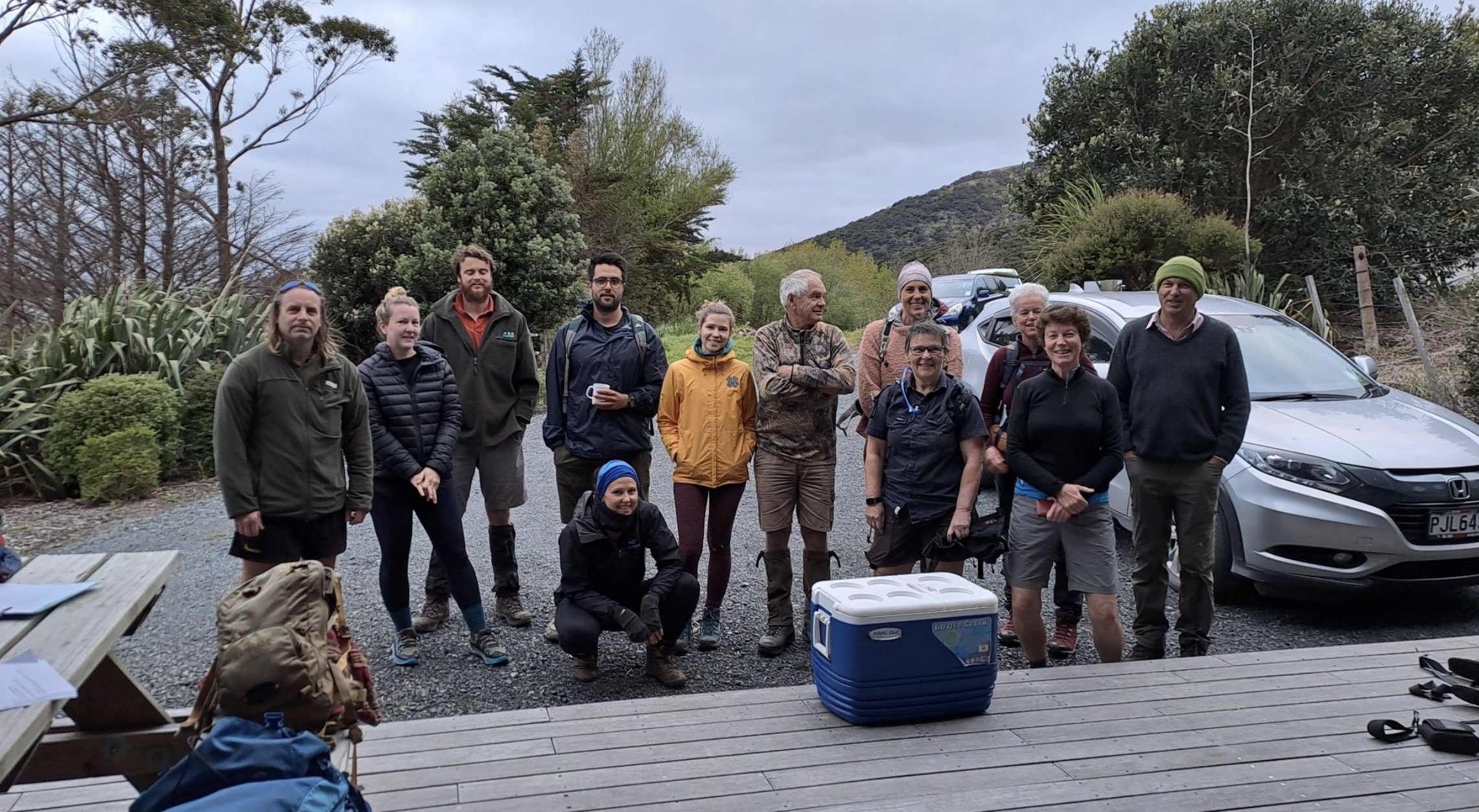 A group of people standing outdoors in front of cars and trees, dressed in casual outdoor clothing, with some holding backpacks and a water bottle, on a cloudy day.