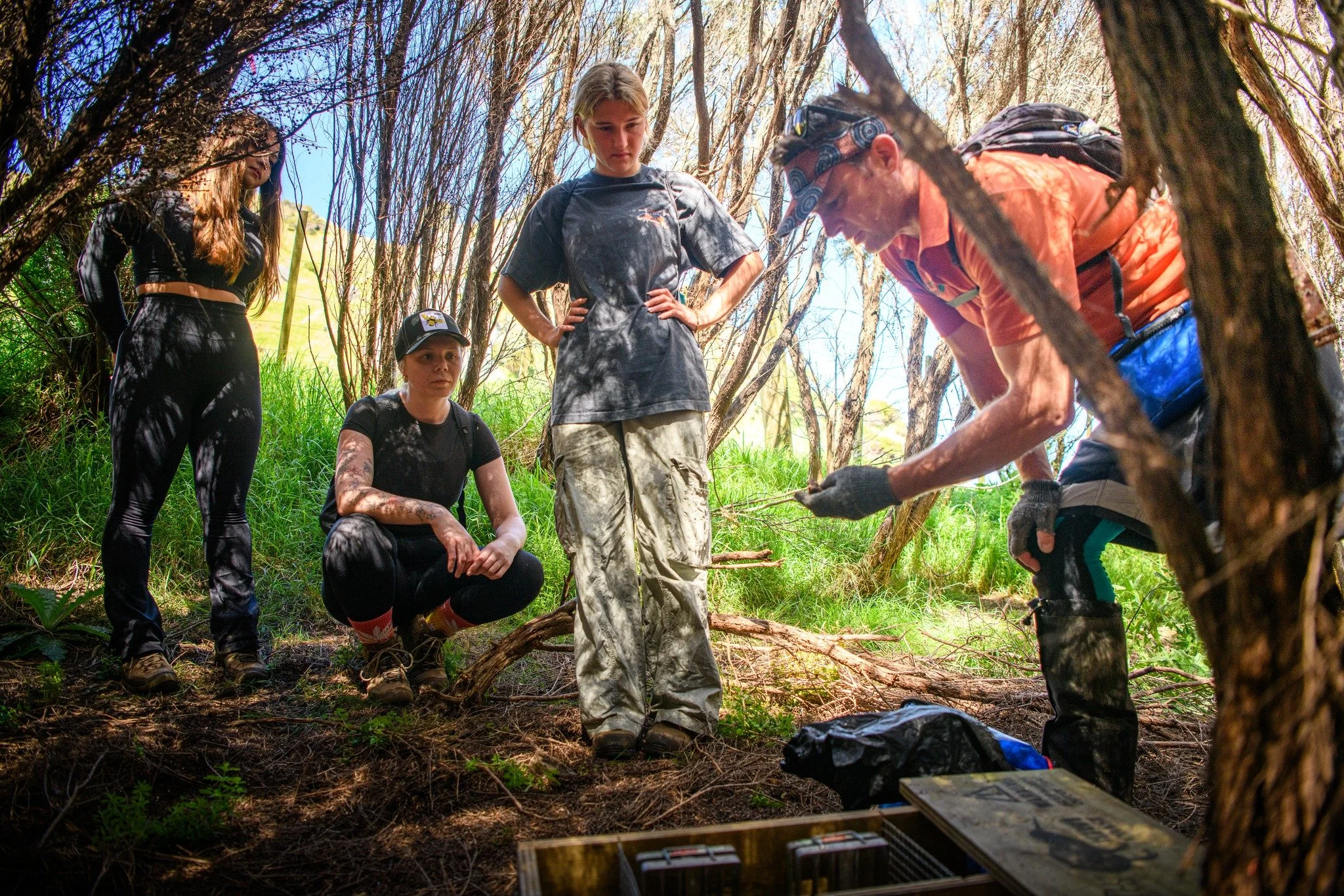 Group of five people in a forest, some sitting and some standing, examining items on the ground; sunlight filters through leafless trees.