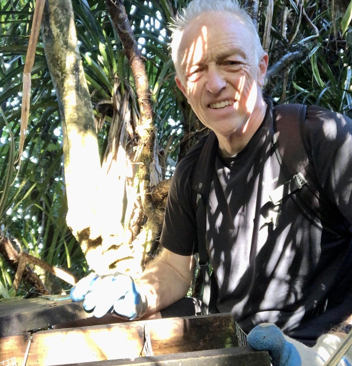A smiling man wearing a black shirt and gloves, with a backpack, in a lush green jungle environment, sitting next to a wooden box or table.