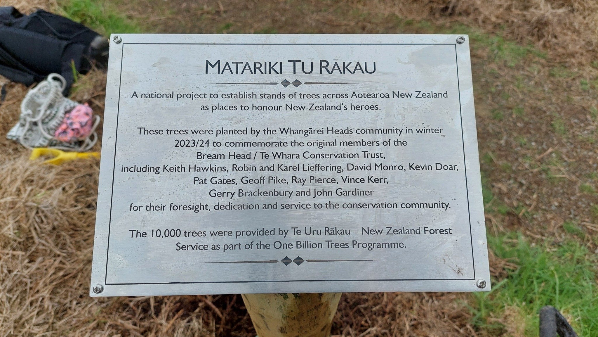 A metal sign on a wooden post describing the Matariki tu Rākau project, including information about tree planting by the Whangārei Heads community in winter 2023/24 to honor New Zealand's heroes, with some objects and foliage visible in the backgroun