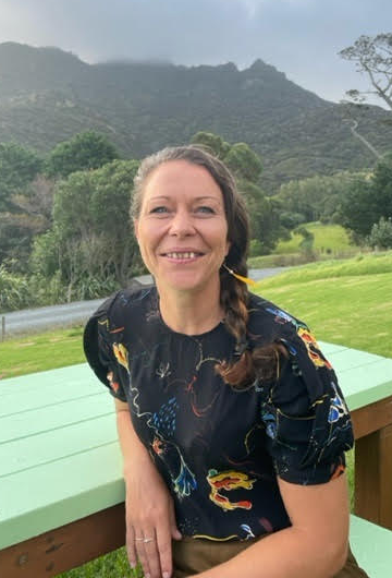 A smiling woman with braided hair standing outdoors near a green wooden table, with lush trees and mountains in the background.