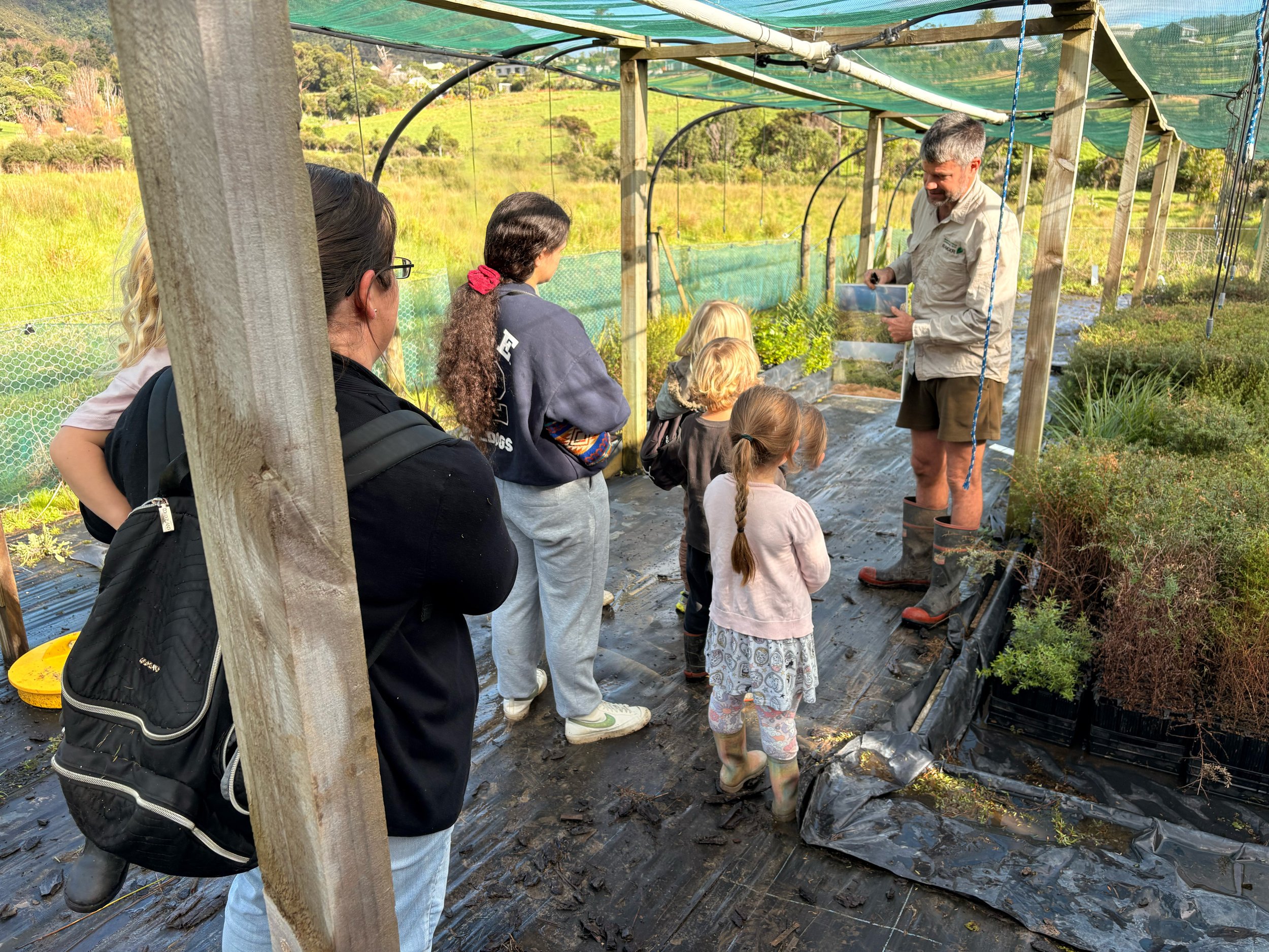 A group of children and adults gathered inside a small greenhouse on a farm, listening to a man explaining something. The greenhouse is covered with green shade cloth, and there are plants and pots around. The background shows a scenic view of rollin