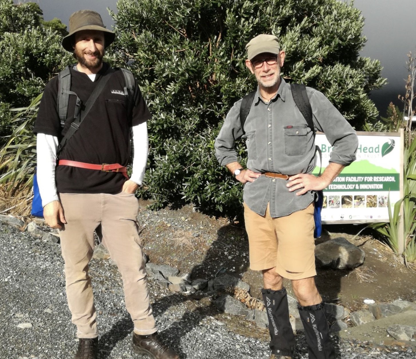 Two men standing outdoors in front of green bushes and a sign, dressed in hiking attire, with backpacks and hats, posing for a photo.
