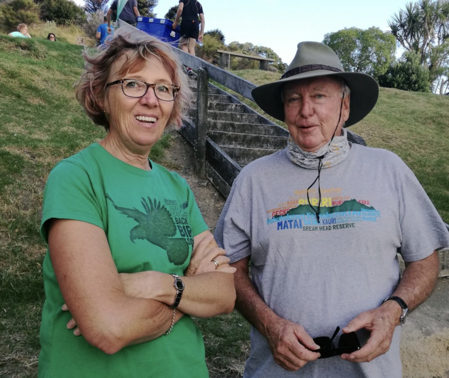 Two people standing outdoors on a grassy hillside with a staircase behind them. The woman on the left is wearing a green T-shirt with a bird graphic, glasses, and has short, wavy, reddish-blonde hair. The older man on the right is wearing a wide-brim