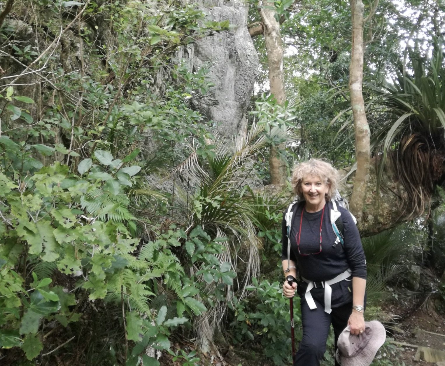 A smiling woman hiking in a lush, green forest with trees and dense foliage, carrying a backpack and holding a walking stick, with a wide-brimmed hat in her hand.