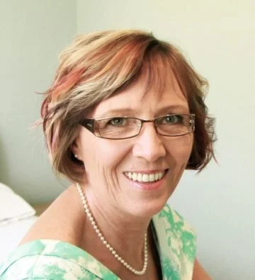 Smiling woman with short, multi-colored hair, wearing glasses, pearl necklace, and a green patterned top.