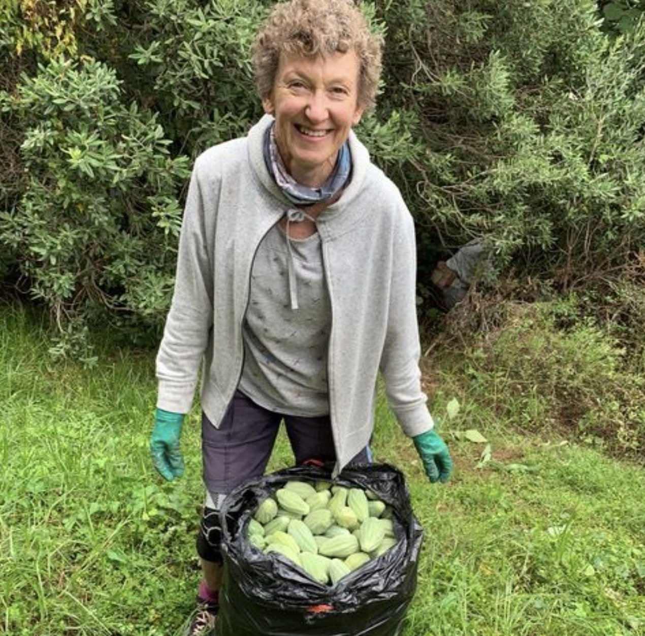 A smiling woman in outdoor clothing, including gloves, standing in a grassy area with greenery in the background, holding a bag filled with harvested green squash.