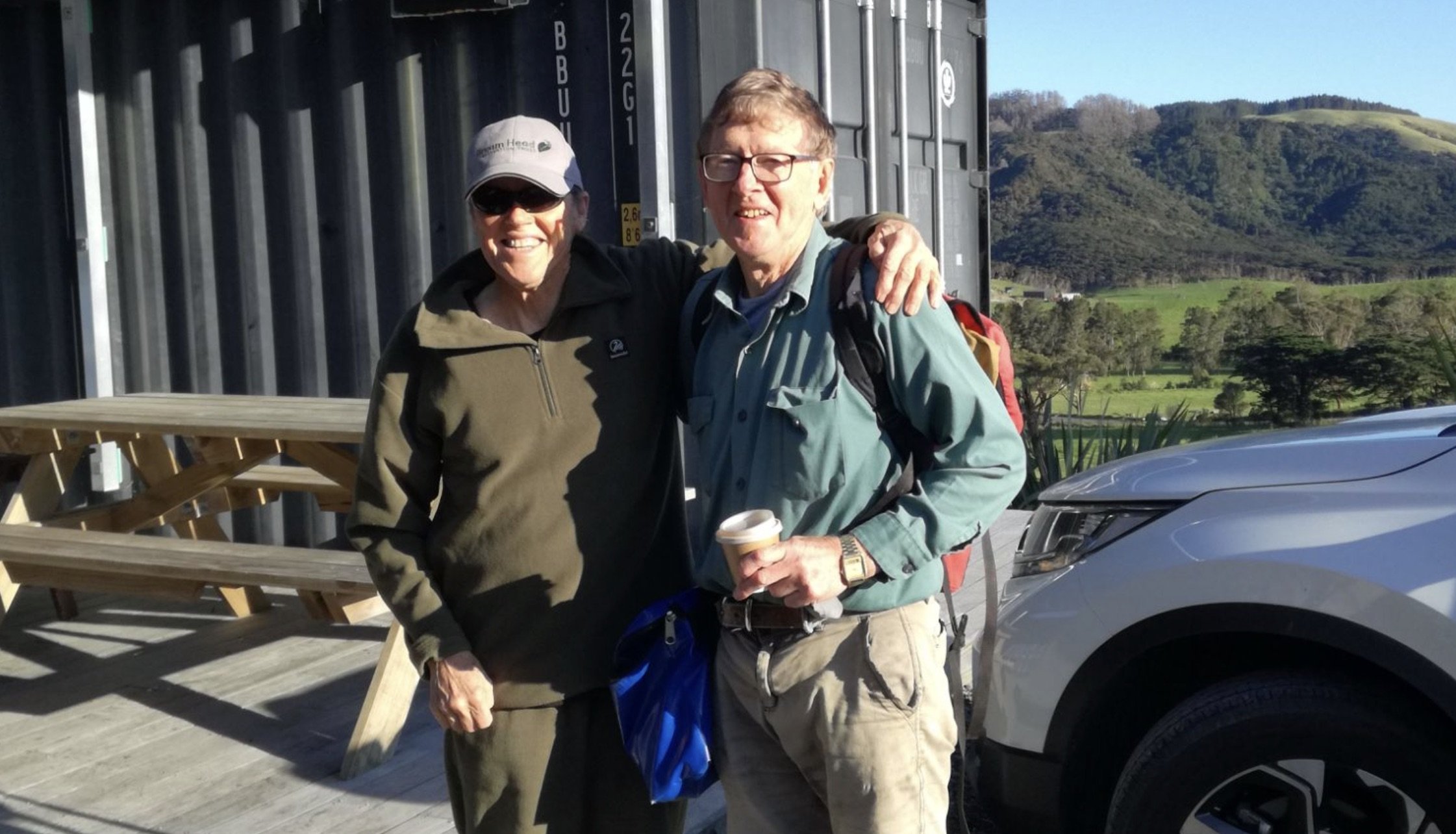 Two people smiling, standing outdoors in front of a gray metal building and a mountainous landscape. One is a woman wearing a cap, sunglasses, and a dark jacket, and the other is a man wearing glasses and a light blue shirt, holding a coffee cup.