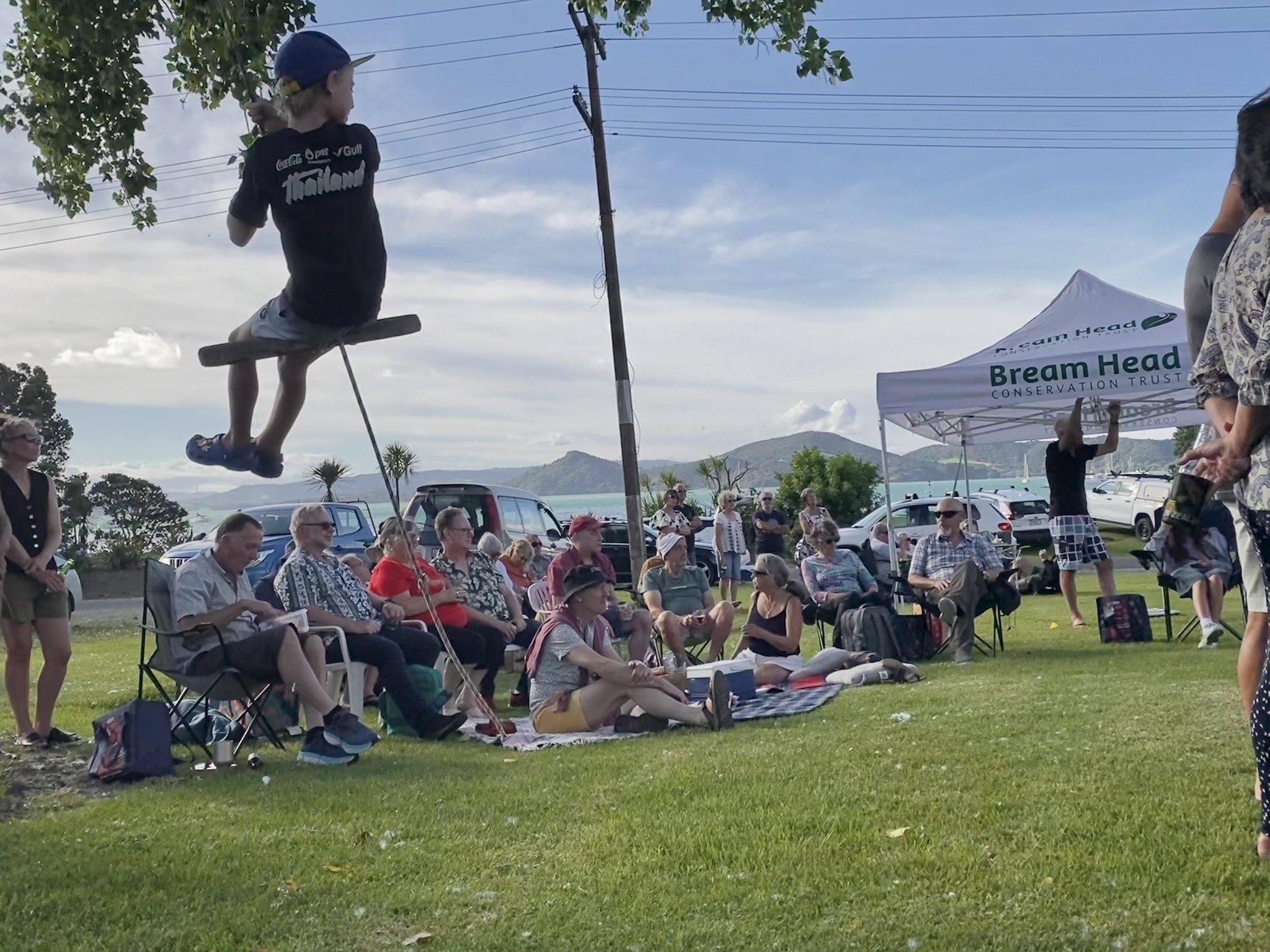 A group of people seated on chairs and a blanket on the grass at an outdoor event, watching a boy performing a tightrope walk between two poles. The event is set against a backdrop of mountains, trees, and a partly cloudy sky, with tents and parked c
