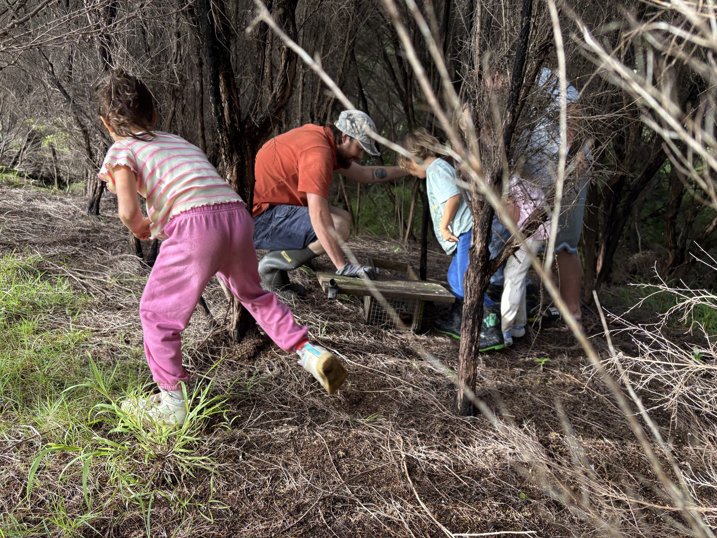 A man with a beard and camouflage cap crouching among bushes with children, planting or working the soil, outdoors in a wooded area.