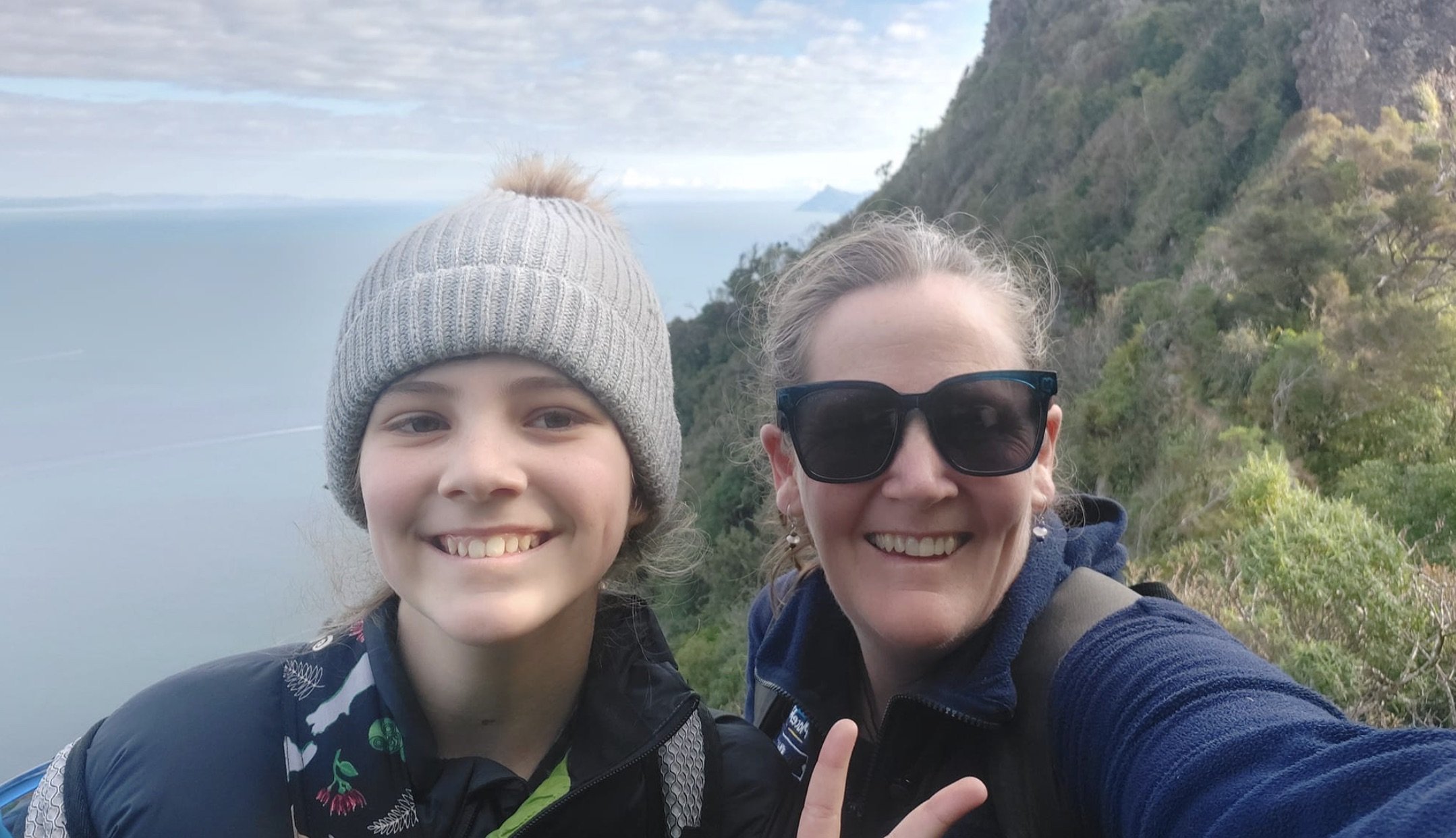 A smiling young person wearing a gray knit hat and a woman with sunglasses taking a selfie together outdoors on a mountain trail. The background features a lush green hillside and a body of water under a partly cloudy sky.