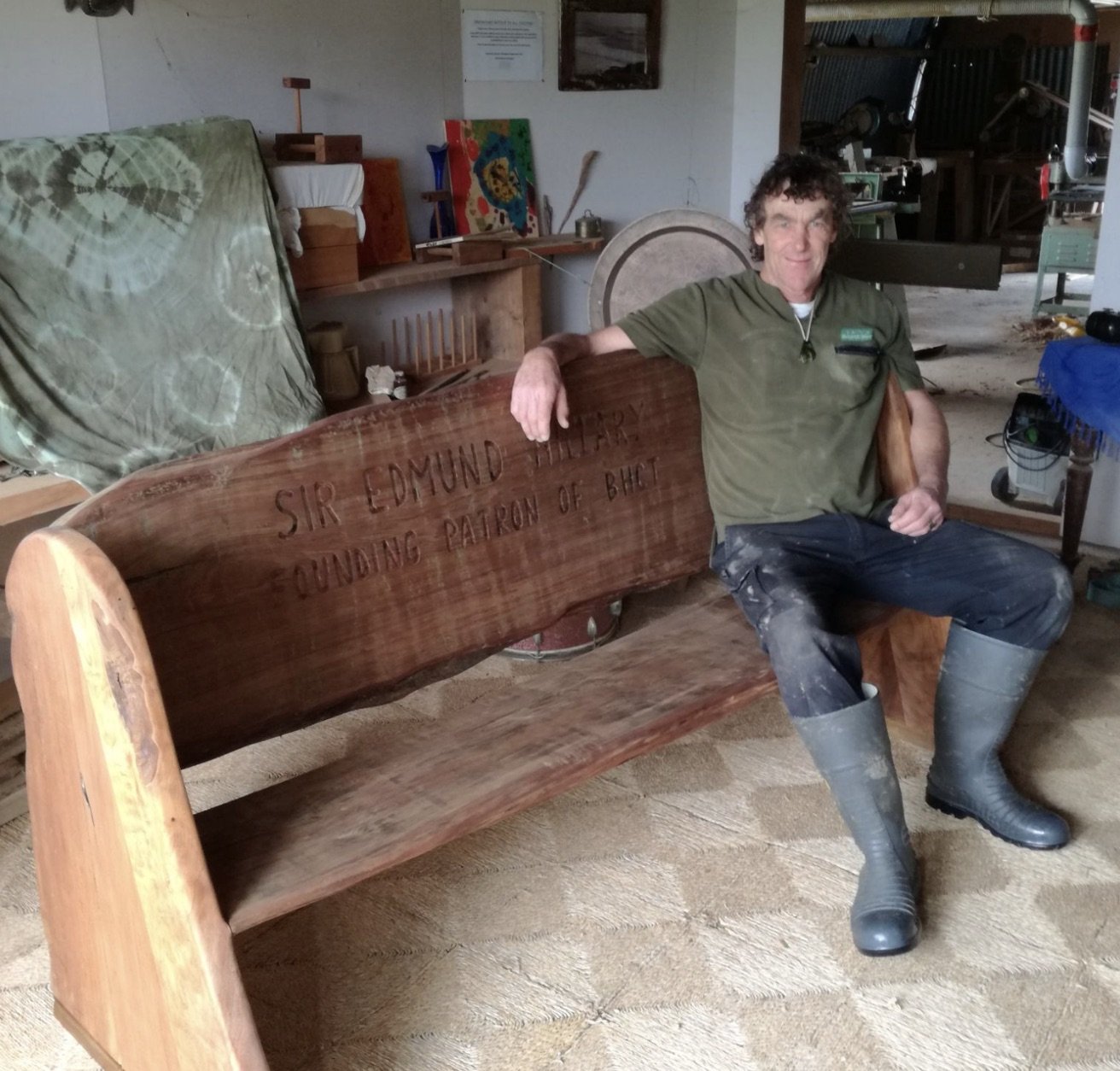 A man sitting on a wooden bench with an inscription that reads, 'Sir Edmund Hillary, founding patron of BHC,' inside a workshop with woodworking tools and paintings in the background.