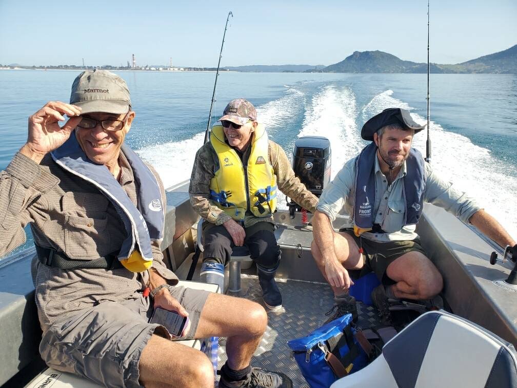 Three men fishing on a boat on a calm body of water with distant mountains and shoreline in the background.