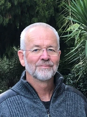 A middle-aged man with glasses, short white hair, and a beard, standing outdoors with green foliage in the background.