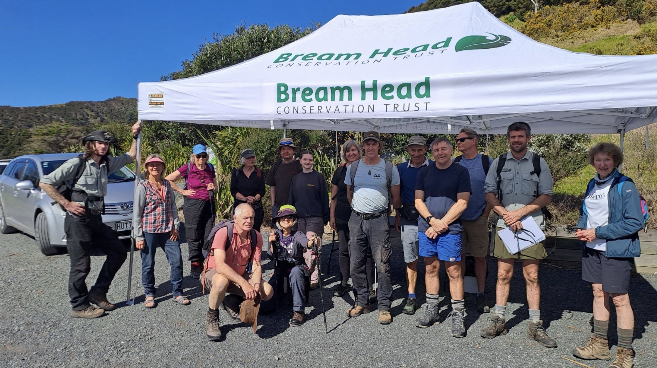 Group of hikers standing under a white tent labeled 'Bream Head Conservation Trust,' with scenic hills and clear blue sky in the background.