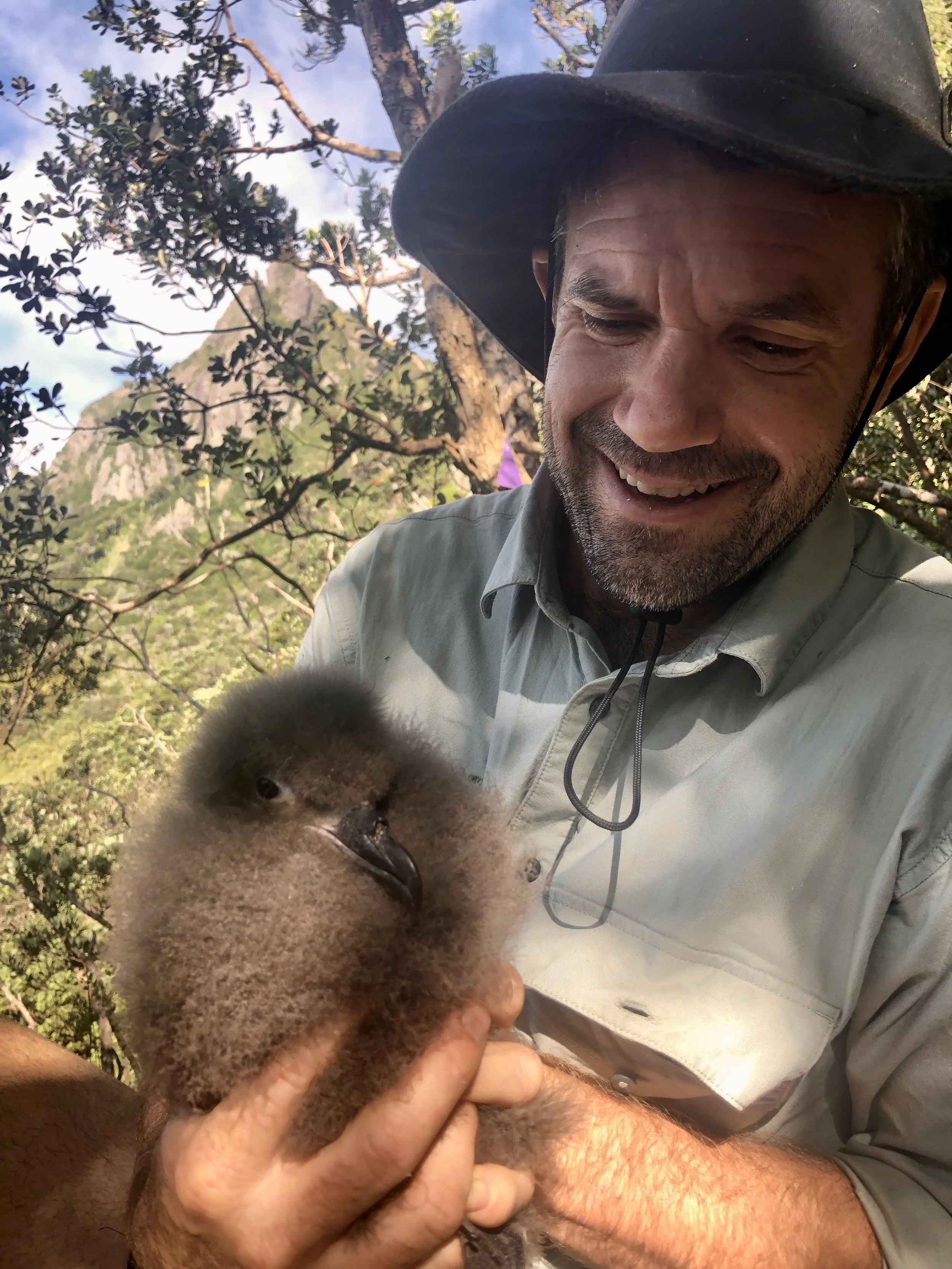 A man wearing a hat and a gray shirt is holding a young duckling with fluffy yellow and brown feathers. The man is smiling and outdoors with trees and a mountain in the background.