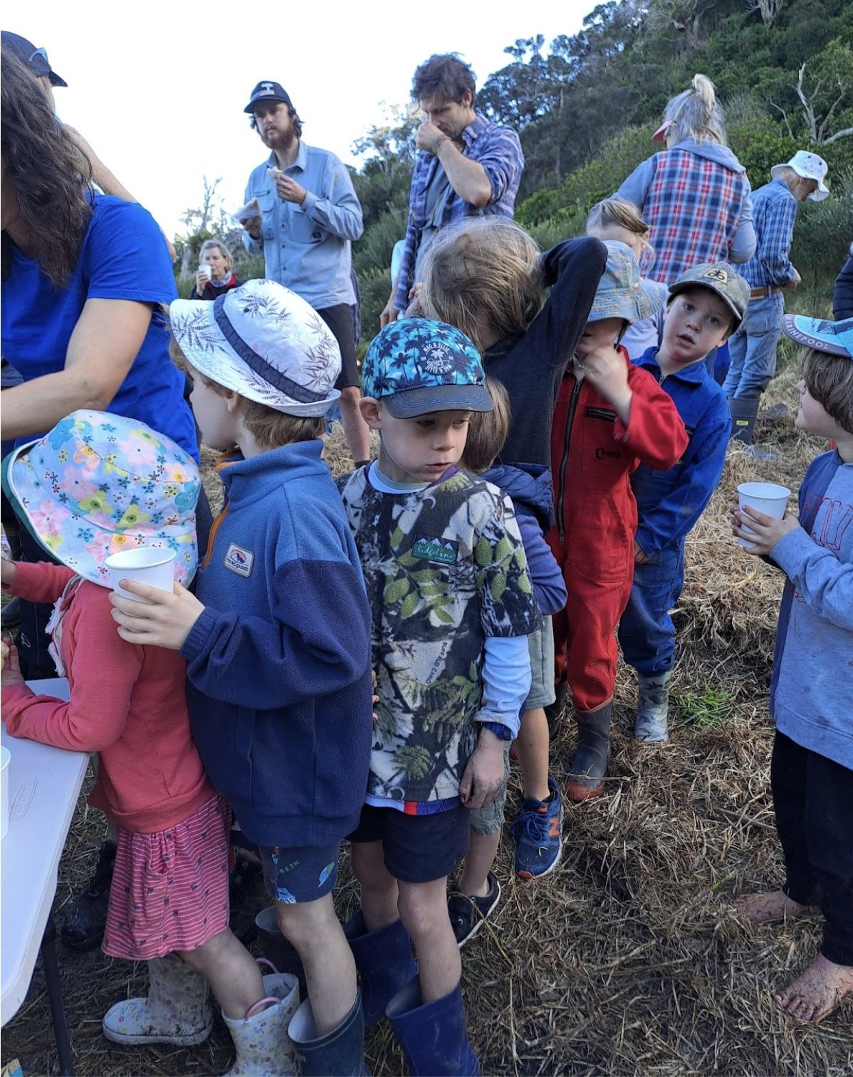 Children and adults gathered outdoors on a farm, some holding cups, with a hillside and trees in the background.
