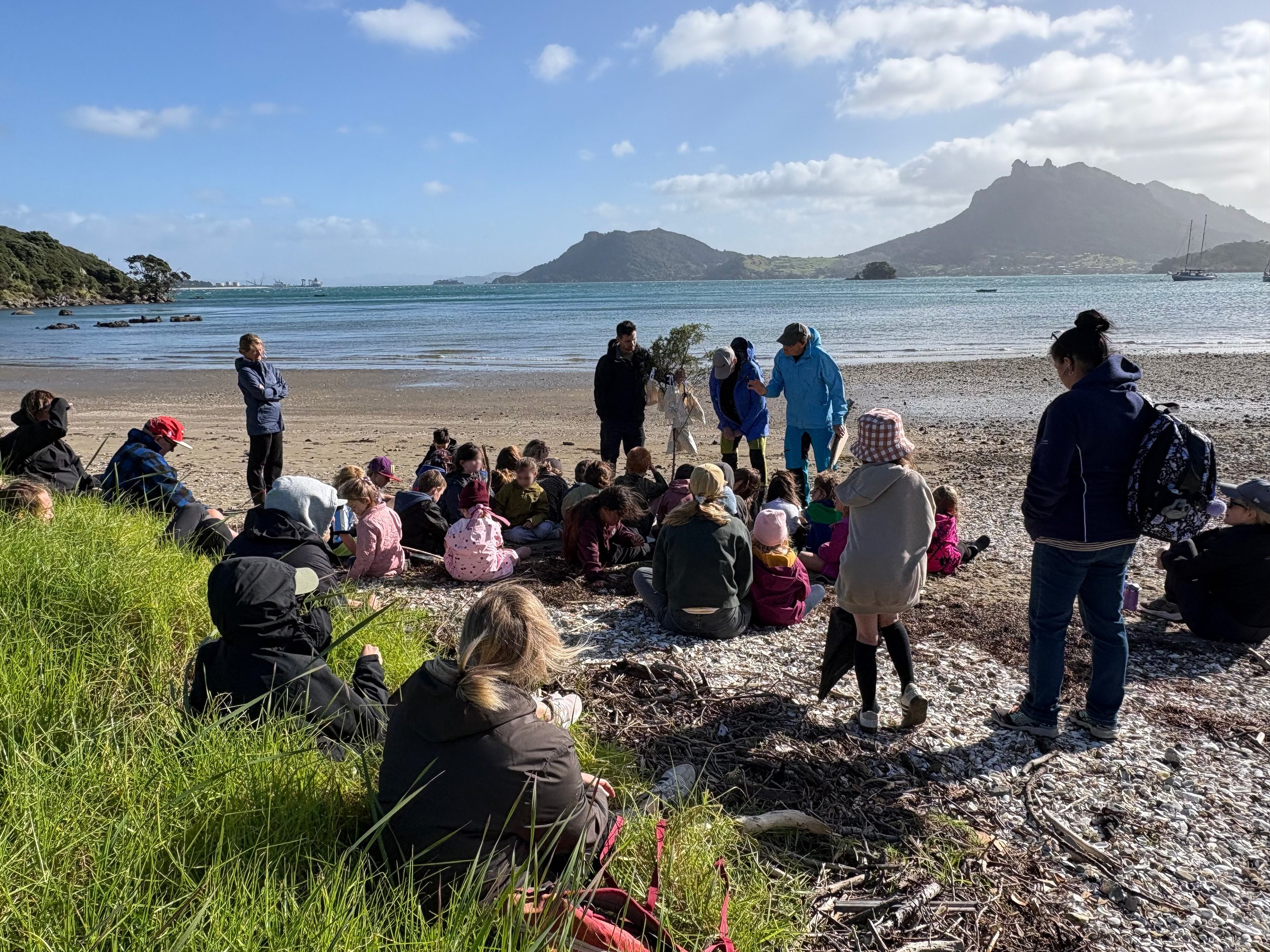 Group of people sitting and standing on a beach near water with mountains and boats in the background under partly cloudy sky.