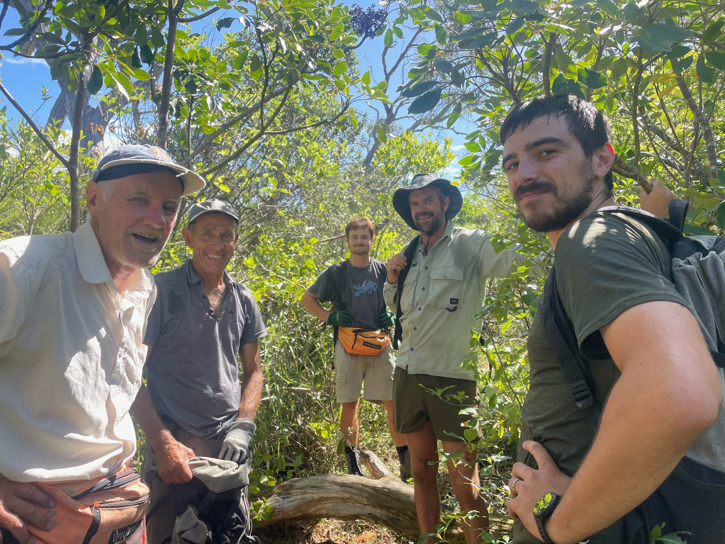 Six men are standing outdoors among trees with green leaves, posing for a group photo. They are smiling and dressed casually, some wearing hats and backpacks, in a sunny environment.
