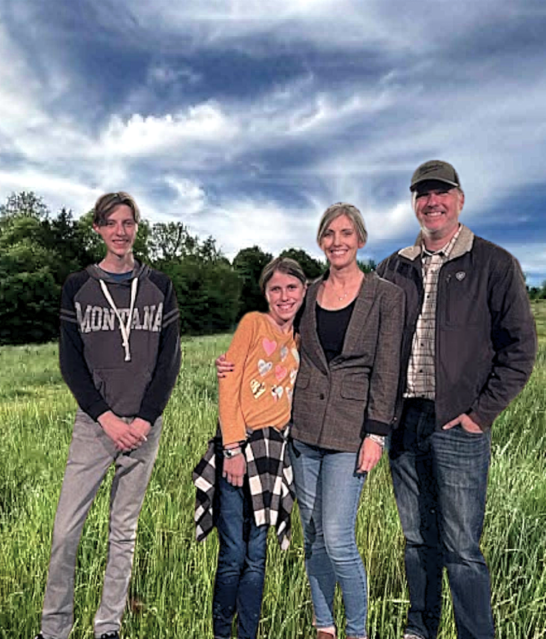 A family of four standing outdoors in a grassy field with trees and a cloudy sky behind them. They are smiling and embracing each other.